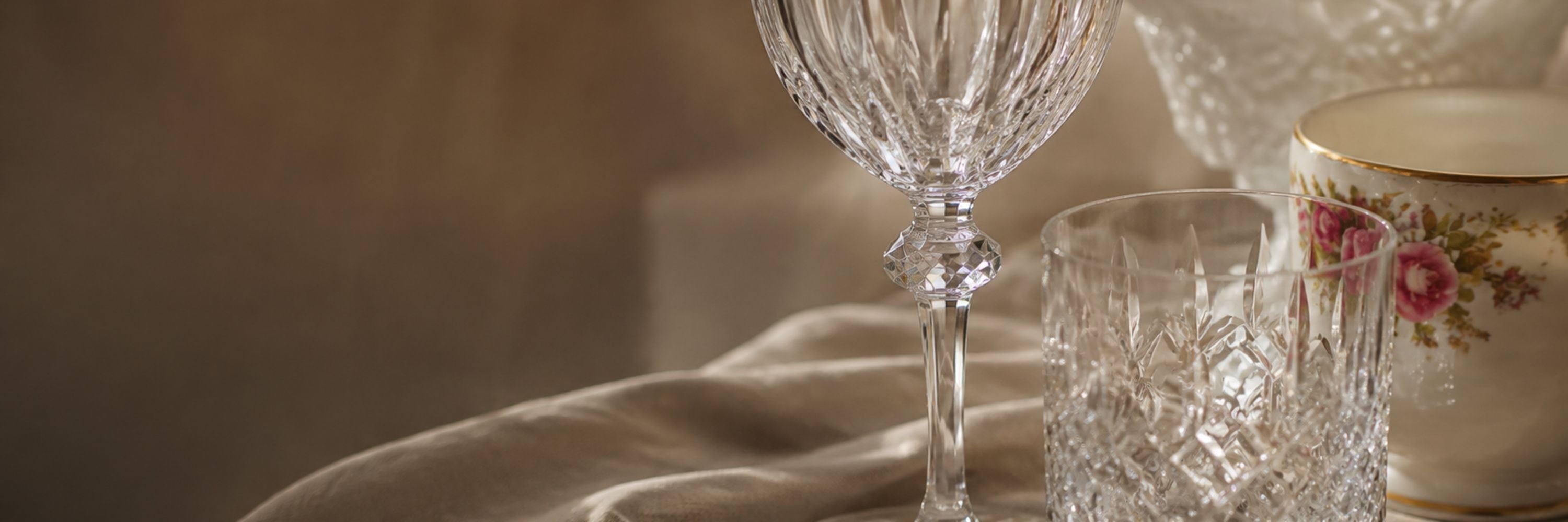 A close-up of clear cut-crystal glassware on draped fabric, including a stemmed wine glass, a tumbler, and part of a cream-colored floral teacup.