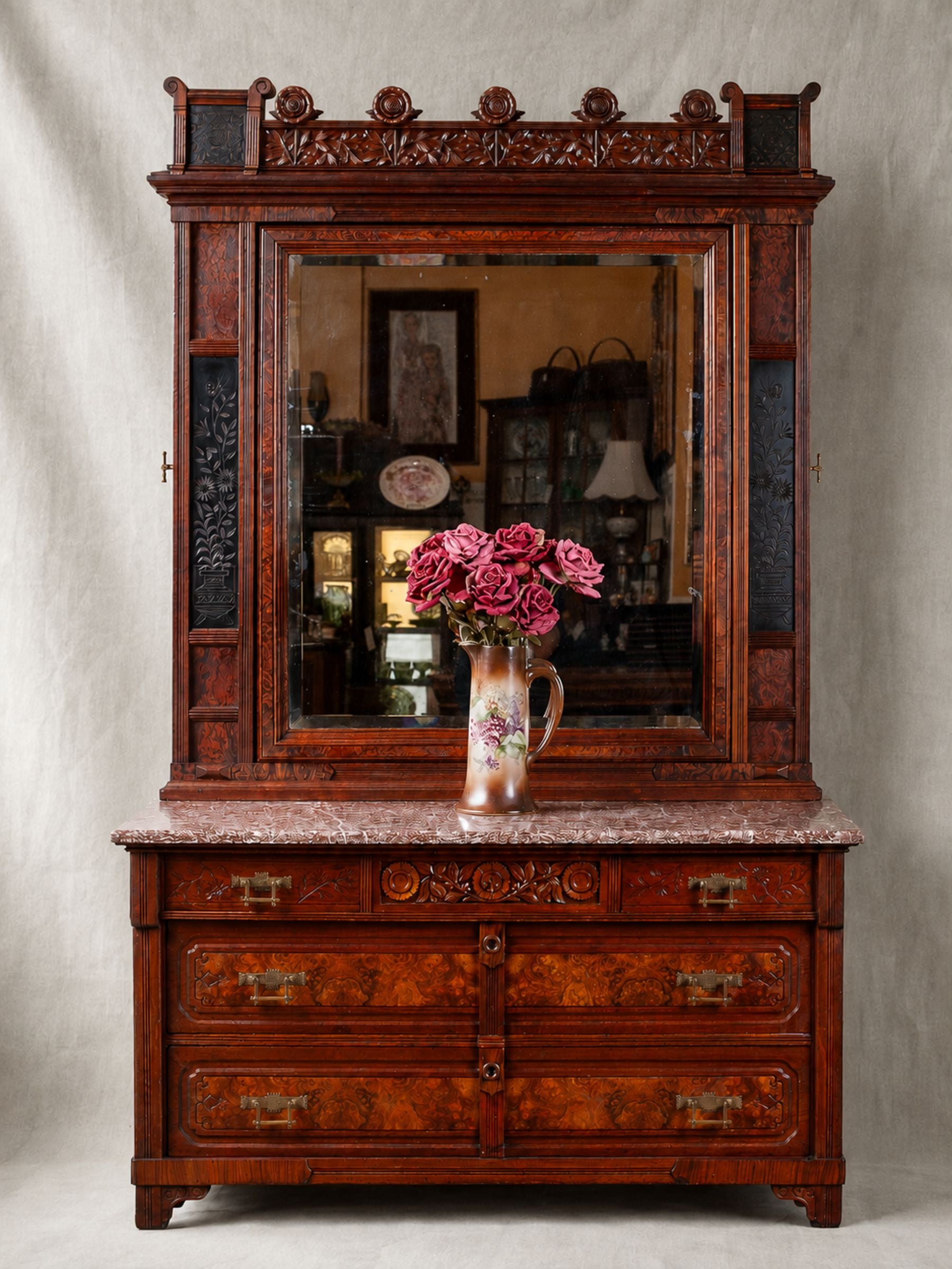 An ornate dark reddish-brown carved wooden hutch with a large mirror, marble-look top, and multiple drawers, styled with a vase of pink roses on top against a neutral studio backdrop.
