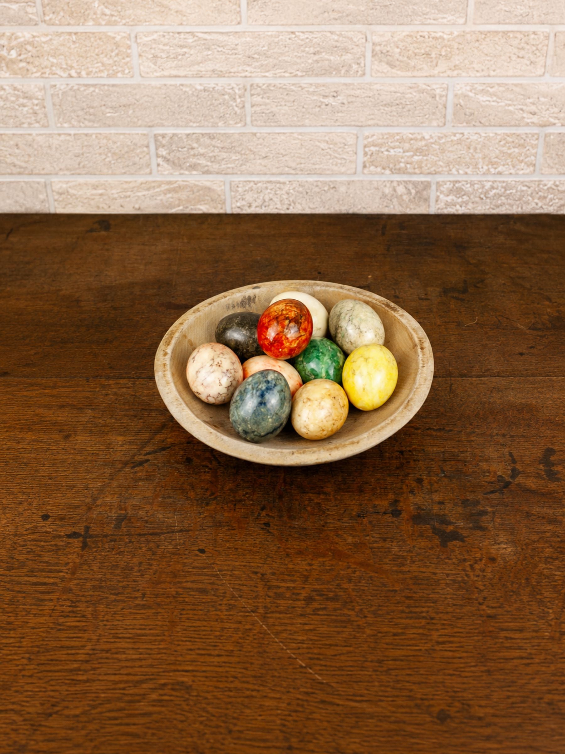 A rustic wooden bowl filled with ten colorful marbleized alabaster eggs, set on a dark brown wooden table against a light beige brick wall.
