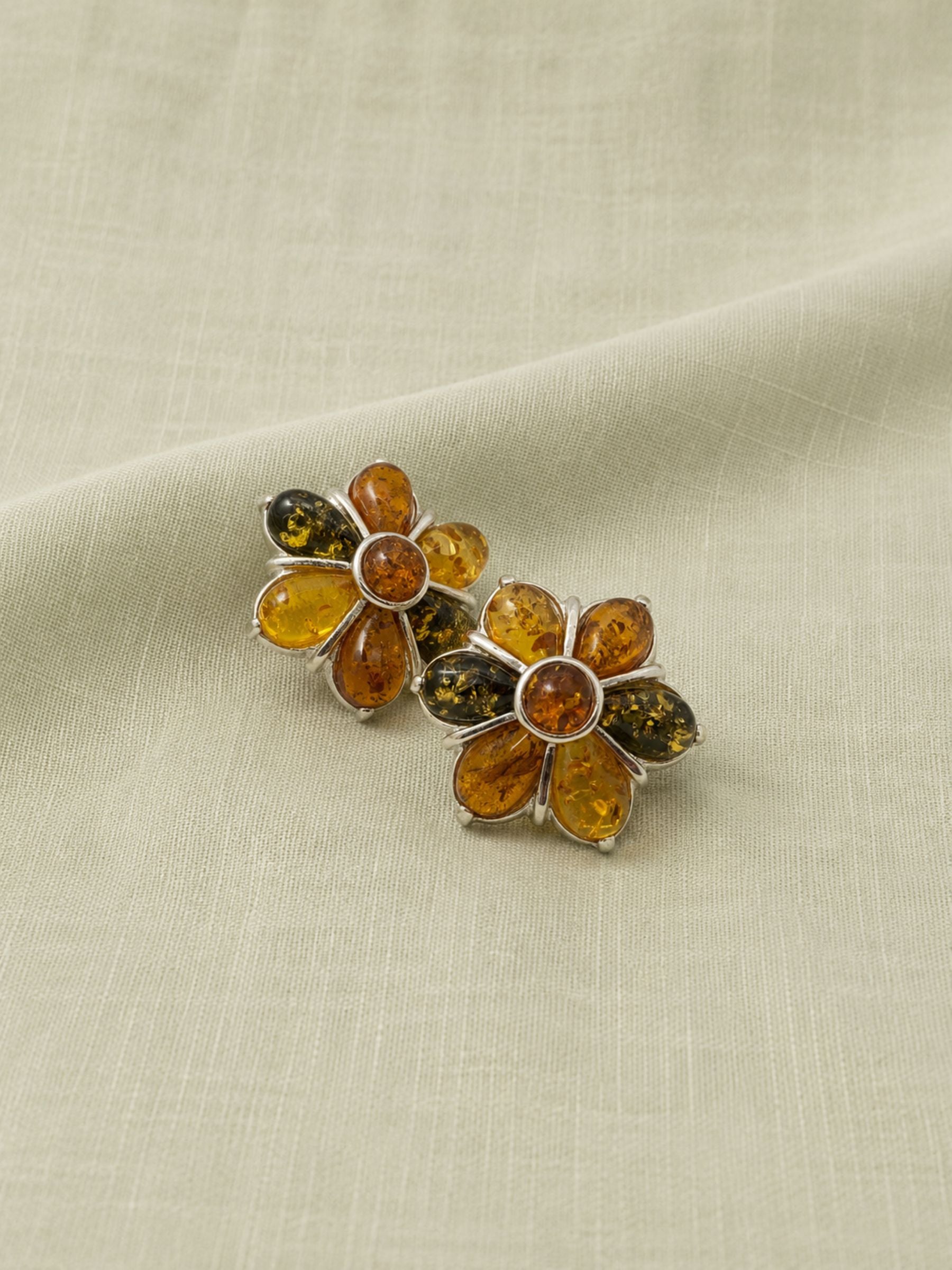 A pair of vintage flower-shaped stud earrings made of genuine amber and sterling silver, displayed on a textured beige fabric background.