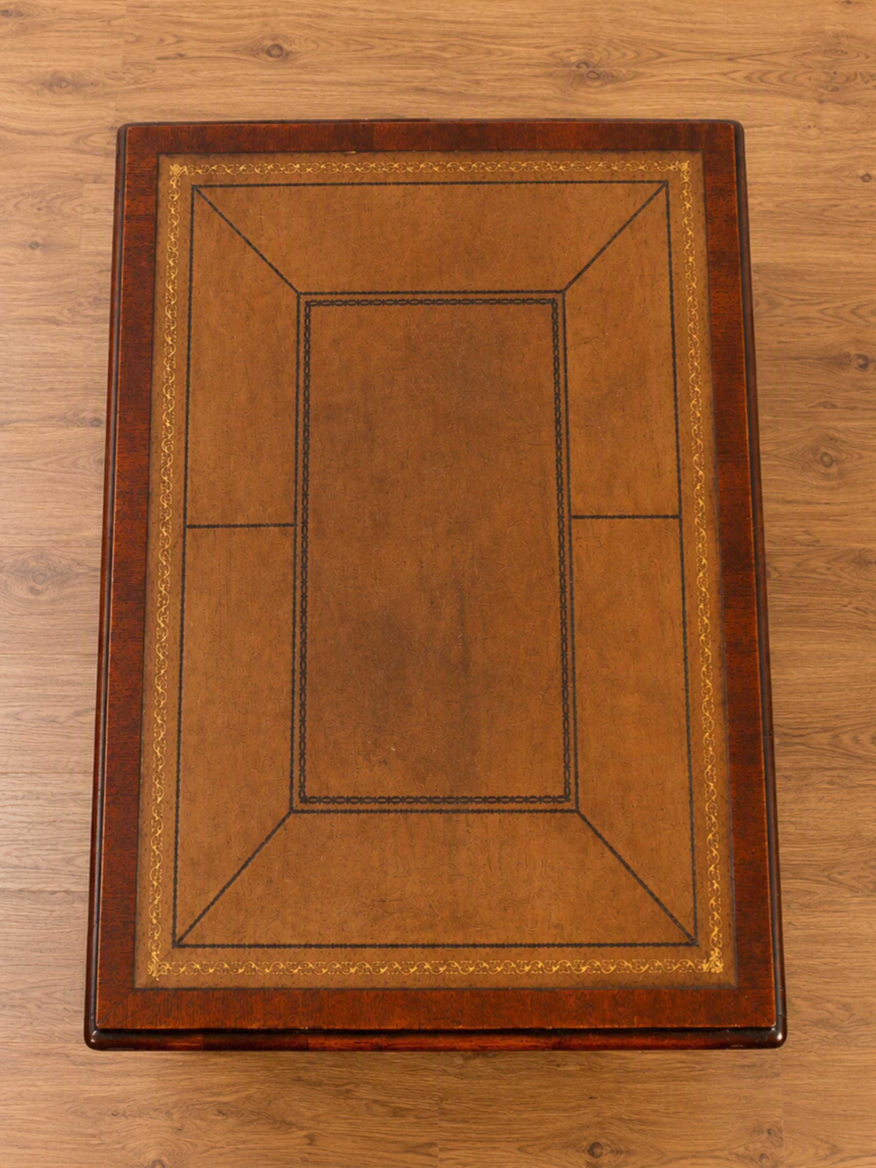 A rectangular wooden table from above, featuring a warm mahogany wood finish with geometric tooled leather details in darker brown and light gold-toned lines, placed on a light wood floor.