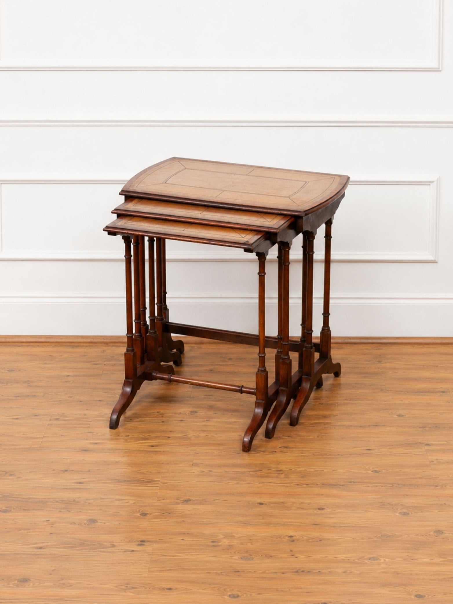 A set of three nested wooden nesting tables with a warm medium-brown finish, standing on a light wood floor against a white paneled wall.
