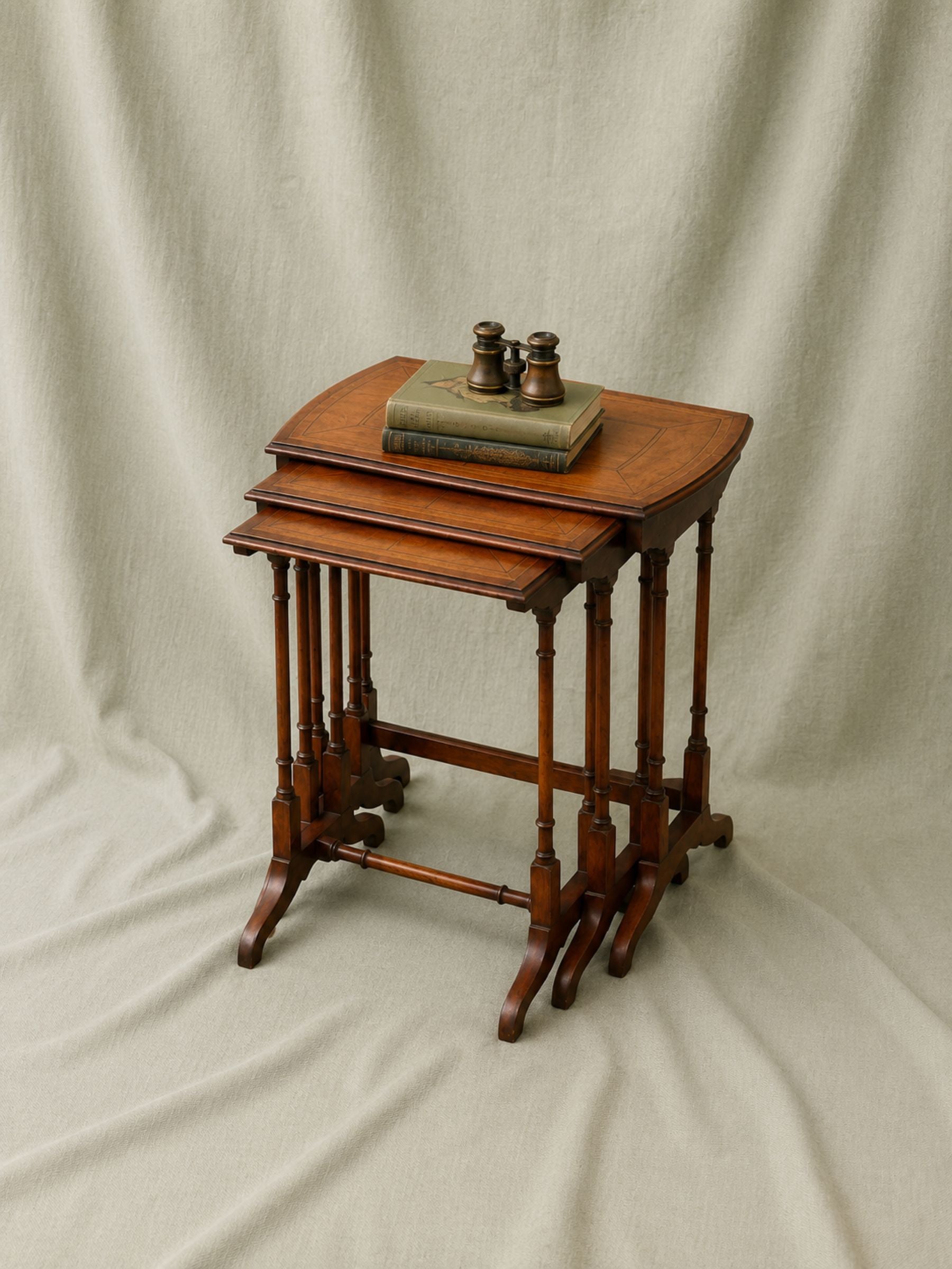 A set of three nested wooden nesting tables with a warm mahogany finish, styled with a vintage book and brass binoculars on top, set against a draped beige fabric backdrop.