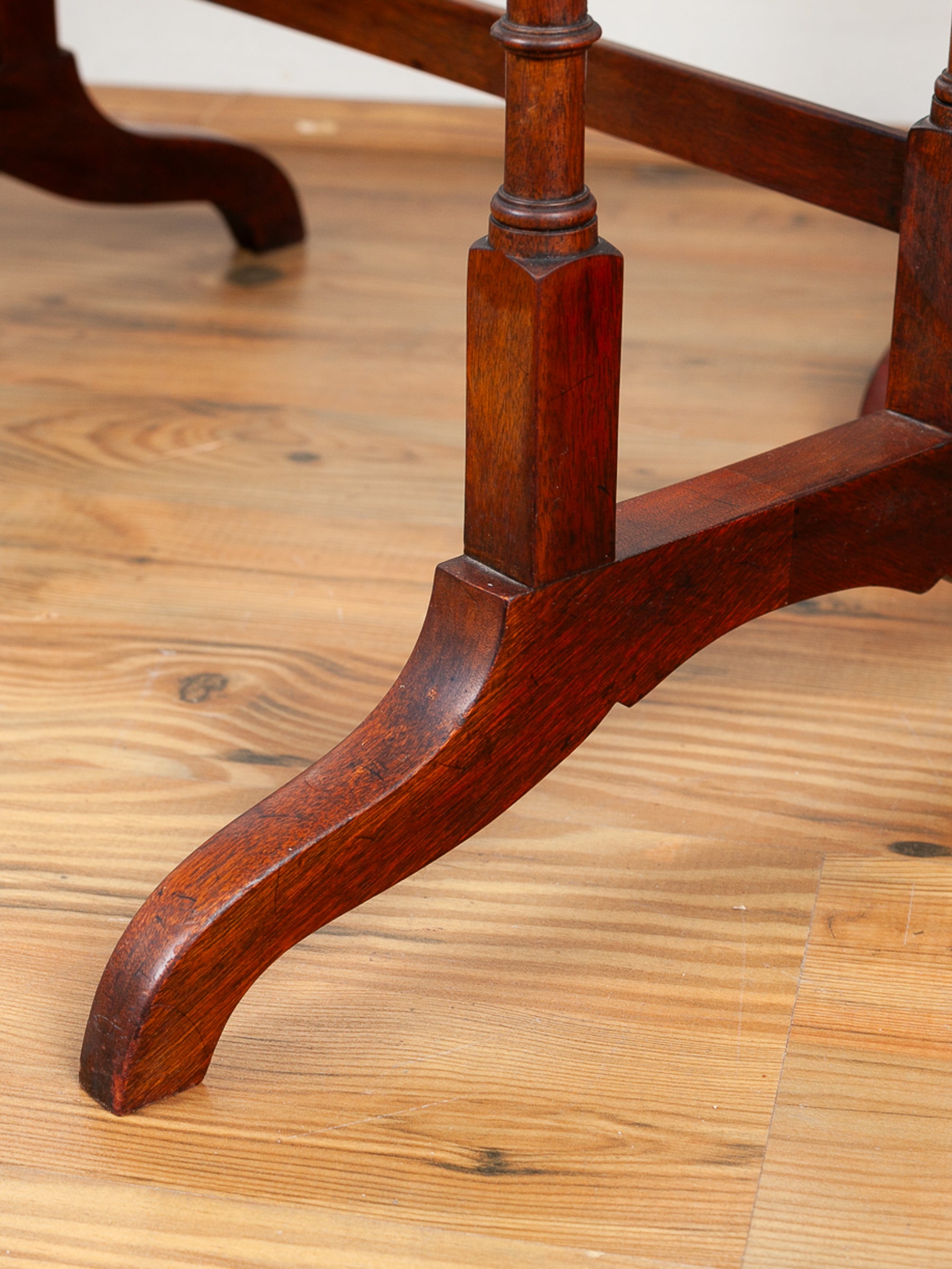 Close-up of a dark reddish-brown wooden table leg with a curved foot and turned joint, resting on a light wood plank floor.