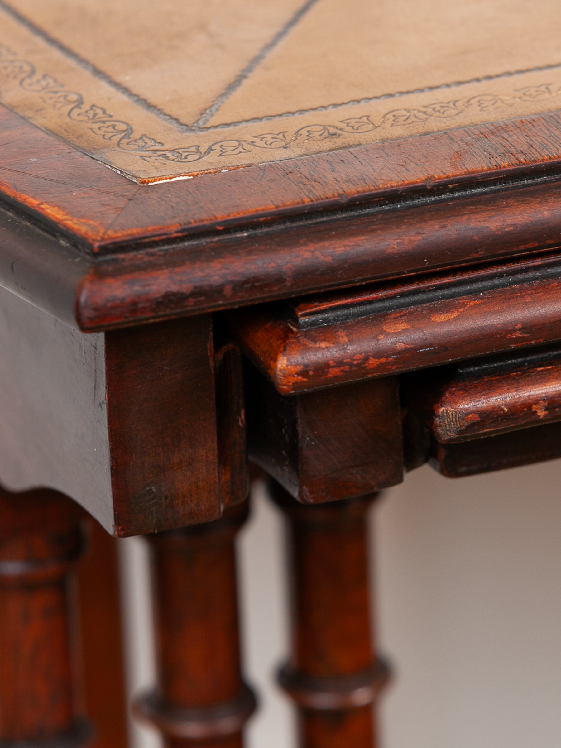 Close-up detail of a dark mahogany wooden table corner, showing carved joinery, turned legs, and part of an olive green leather tabletop with a decorative border pattern.