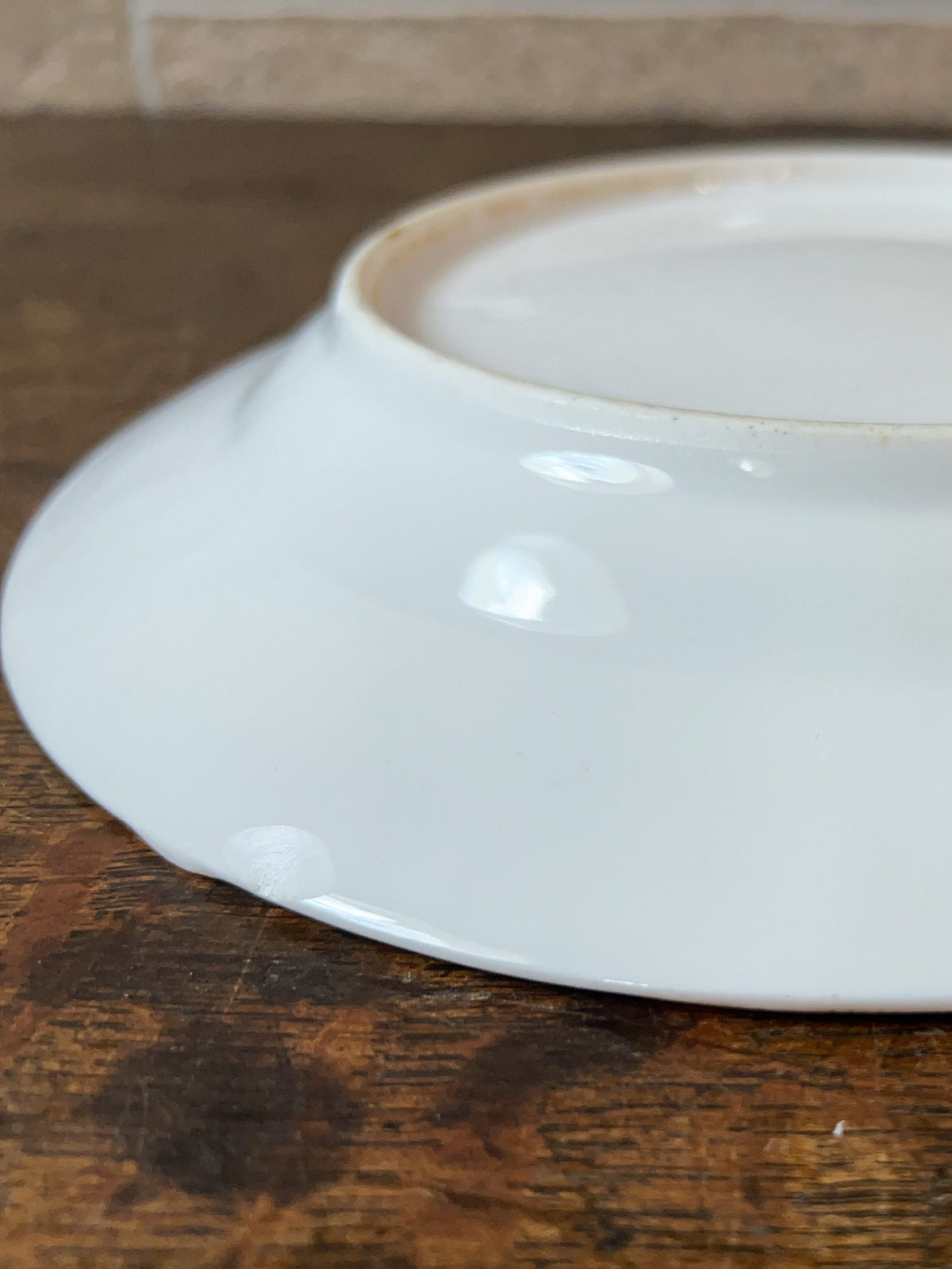 A white ceramic plate shown from the bottom, displaying its glossy glaze and rim, resting on a dark wooden tabletop with a tiled backsplash in the background.