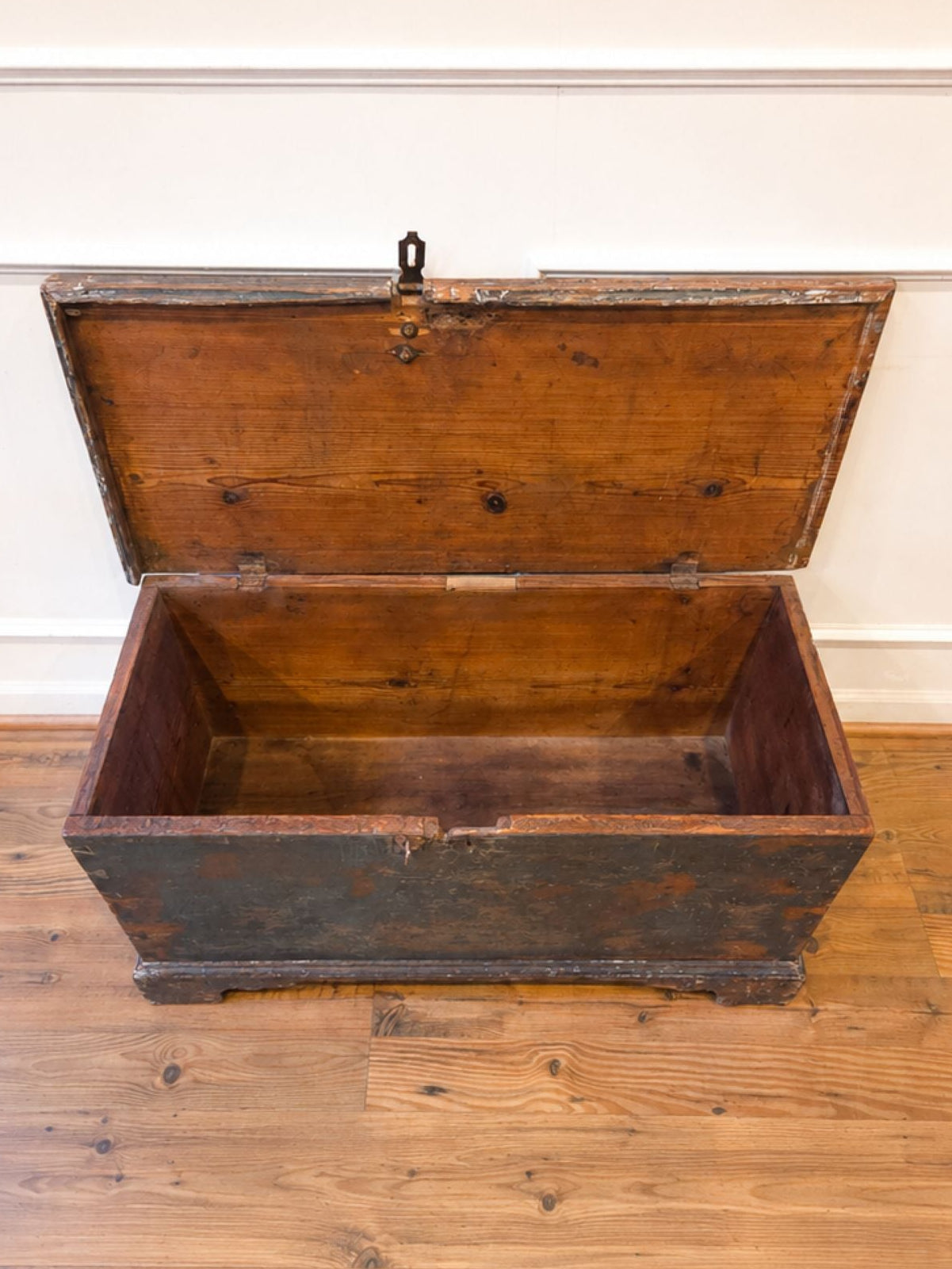 An open vintage wooden chest with a dark weathered exterior and warm brown interior, set on a wooden floor against a white paneled wall.
