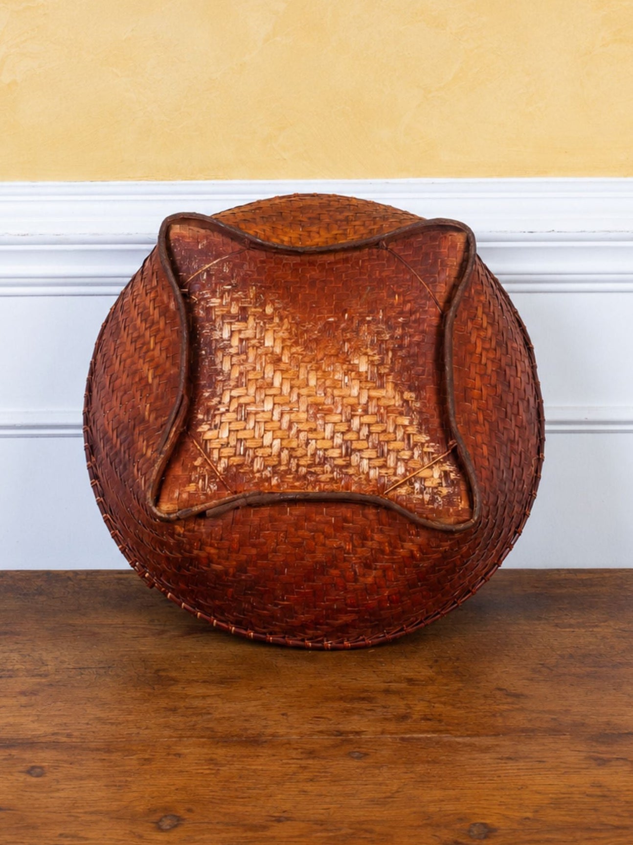 The underside of a round woven rattan or wicker basket displayed on a wooden table against a white chair rail and textured yellow wall.