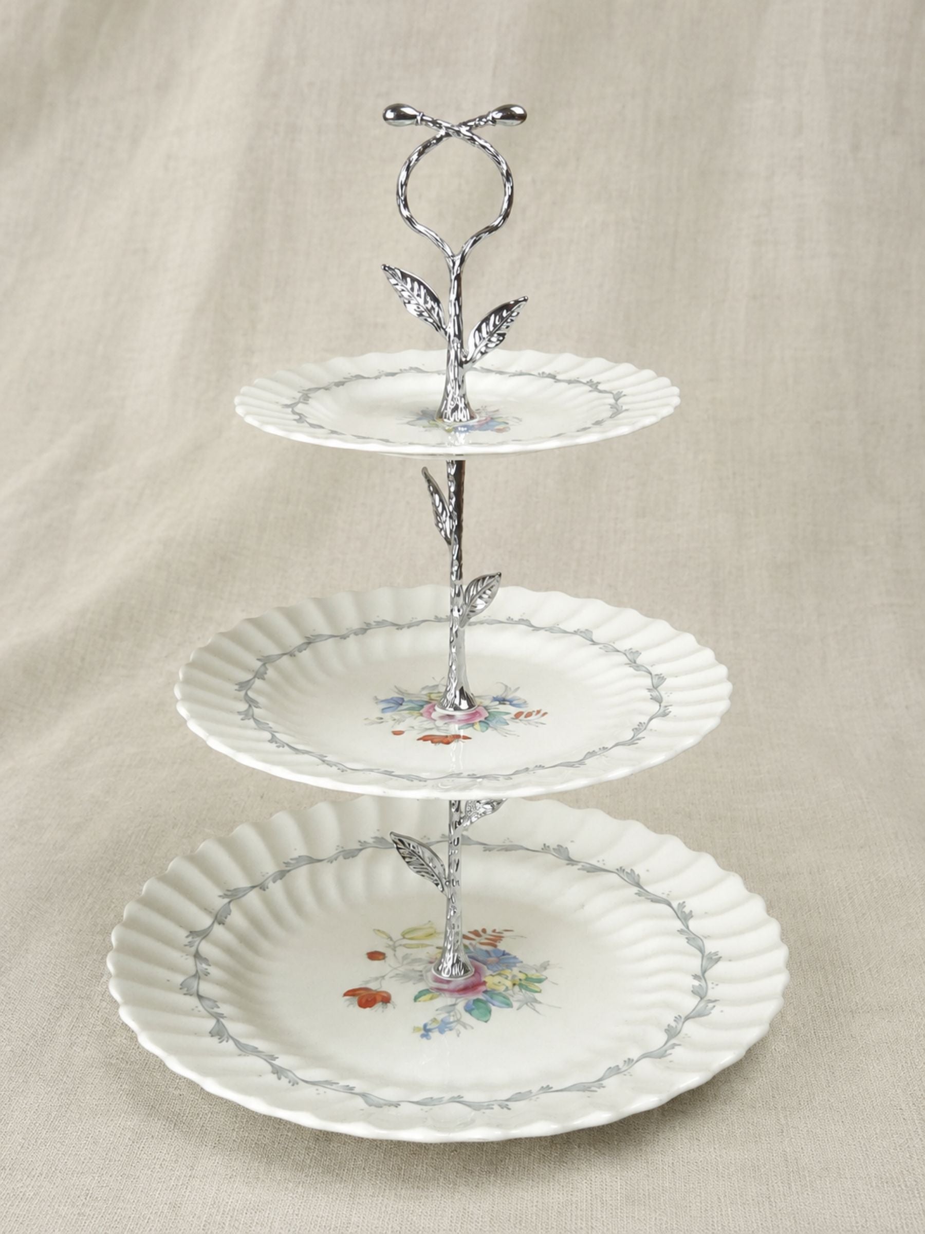 Three-tiered white ceramic serving stand with scalloped plates, floral center designs, and a decorative silver metal vine-style center column on a beige fabric background.