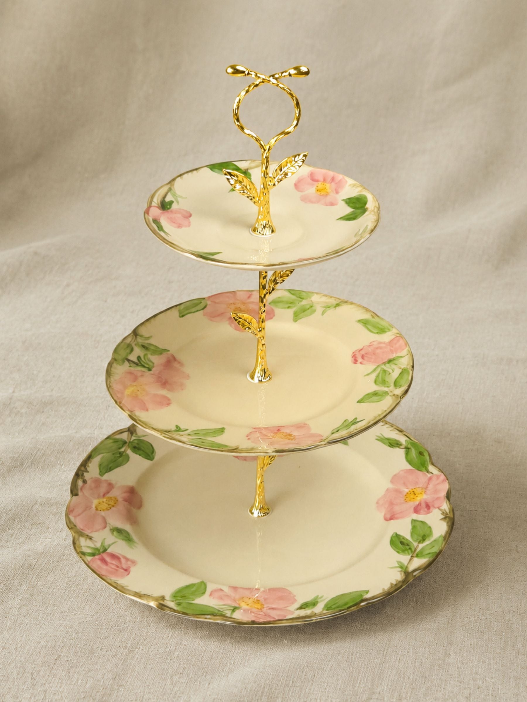 A three-tiered serving stand with a gold metallic trellis-style center and handle, decorated with pink flowers and green leaves on a cream-colored ceramic plate, set against a neutral beige fabric backdrop.