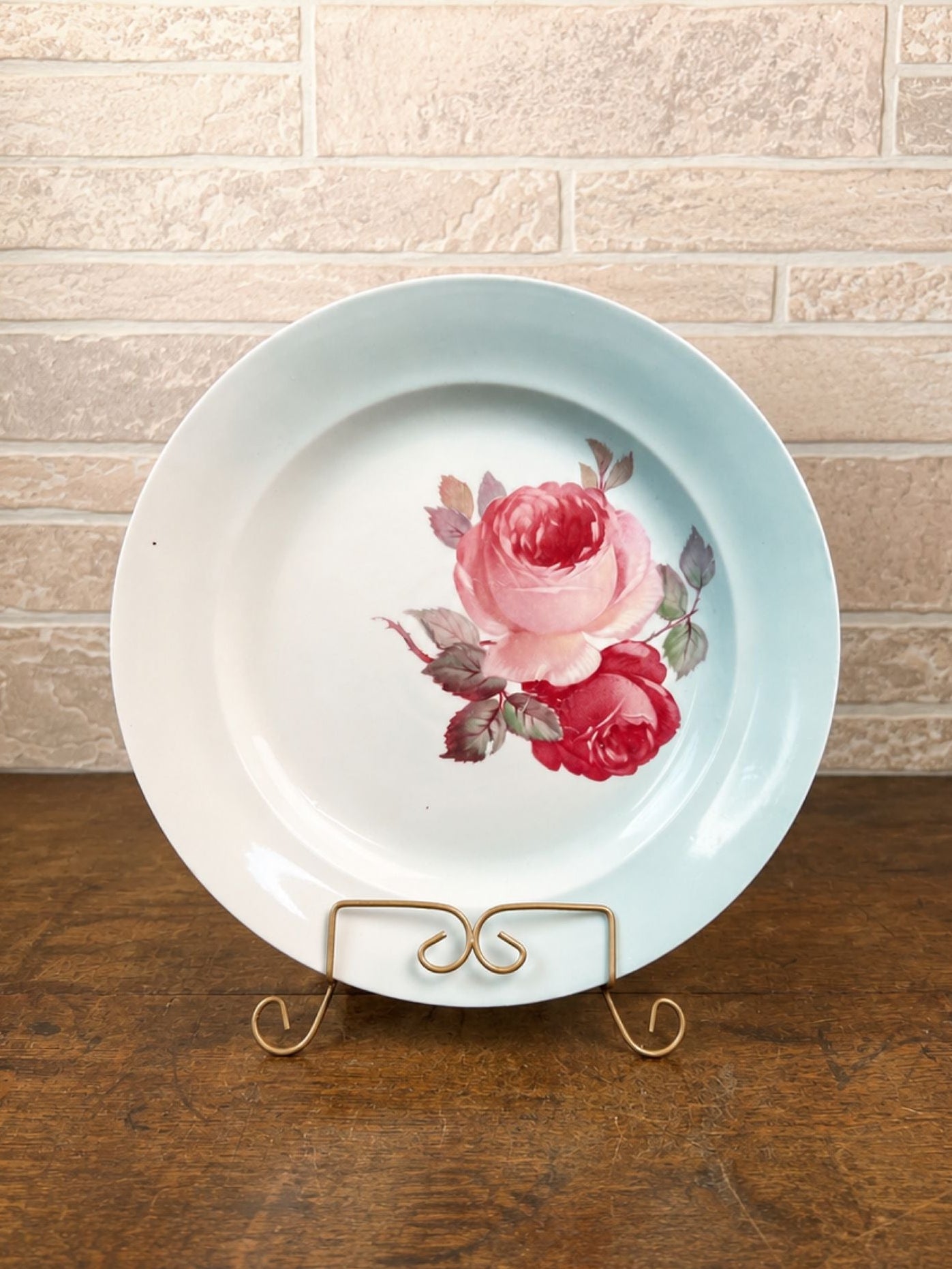 A round white ceramic plate with a light blue rim and a painted pink rose design in the center, displayed on a small gold wire stand on a wooden table against a light brick wall.
