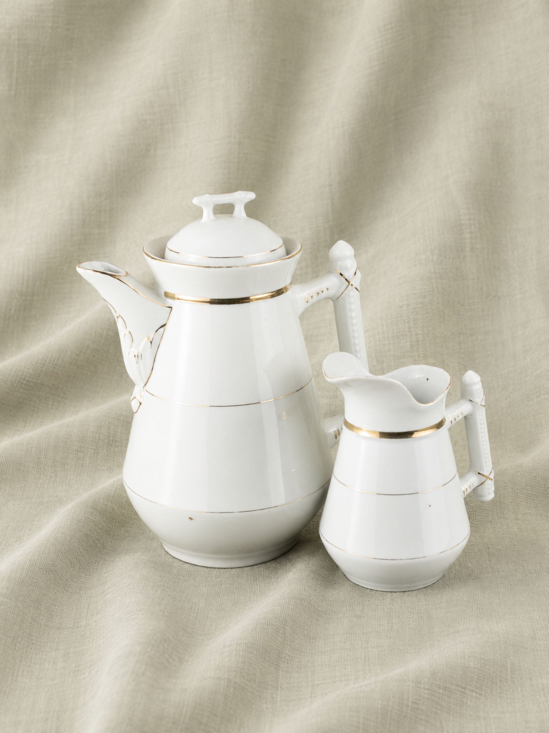 A white porcelain coffee pot and matching creamer pitcher with thin gold trim, set against a draped beige linen fabric background.