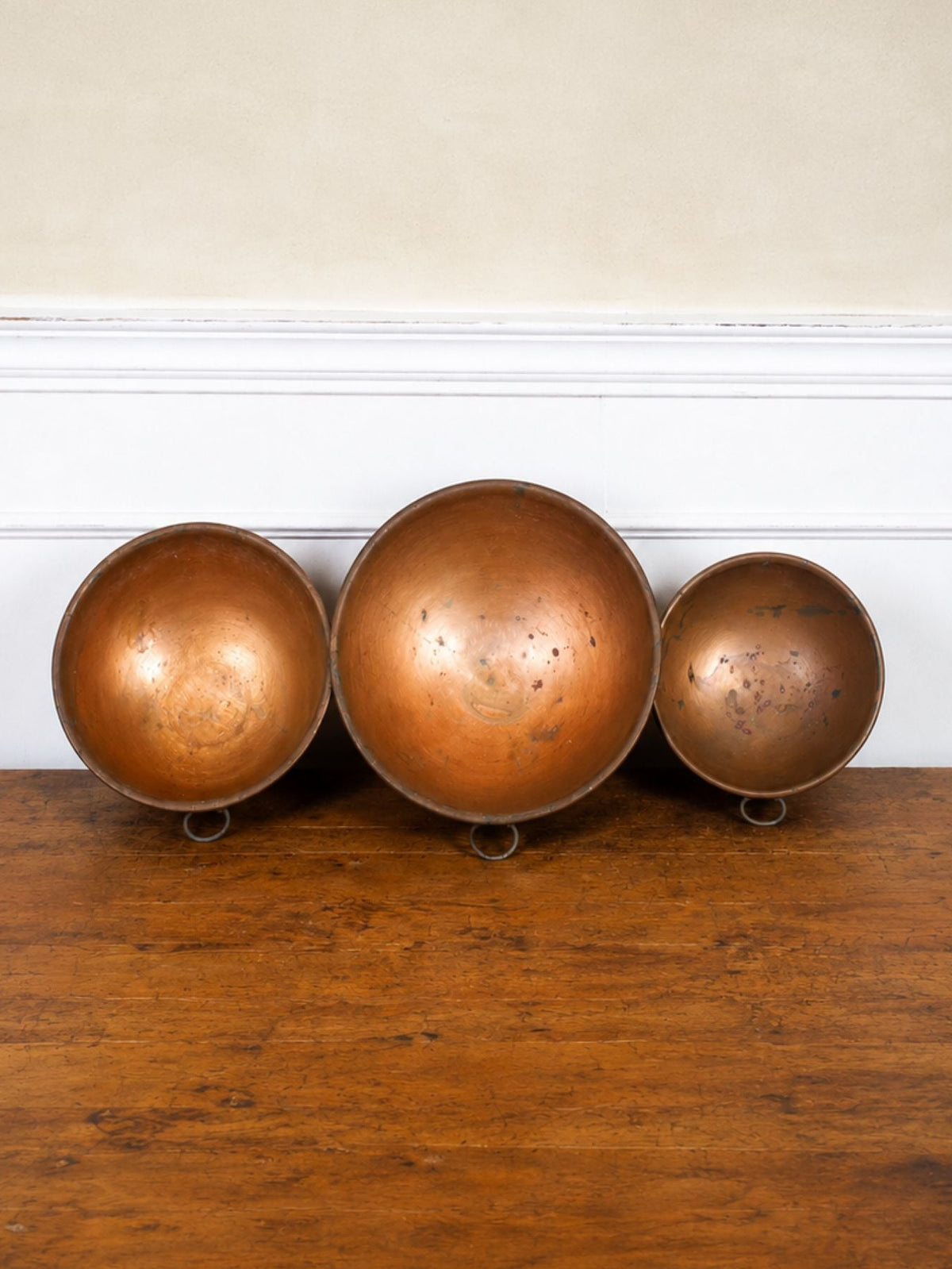 Three vintage copper bowls of varying sizes displayed on a wooden table against a cream wall with white trim.