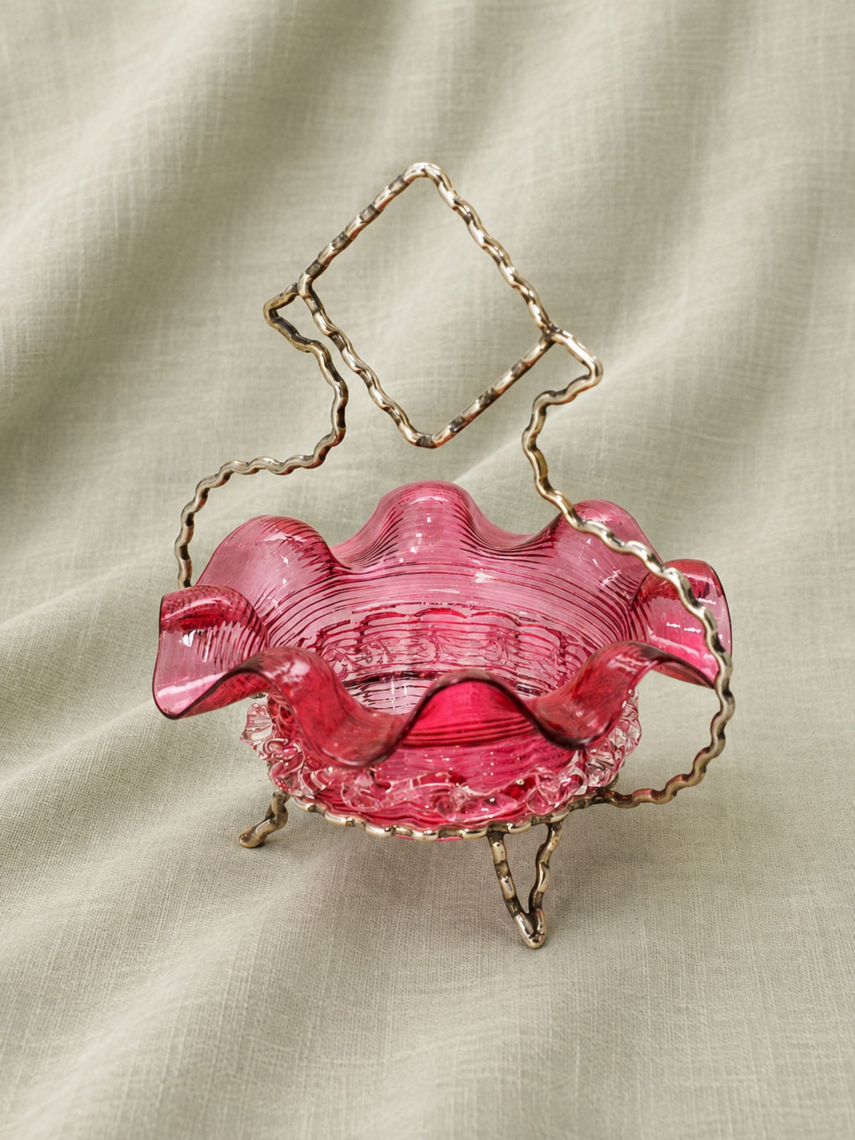 A pink ruffled glass bowl with a decorative silver stand, set against a draped beige fabric background.