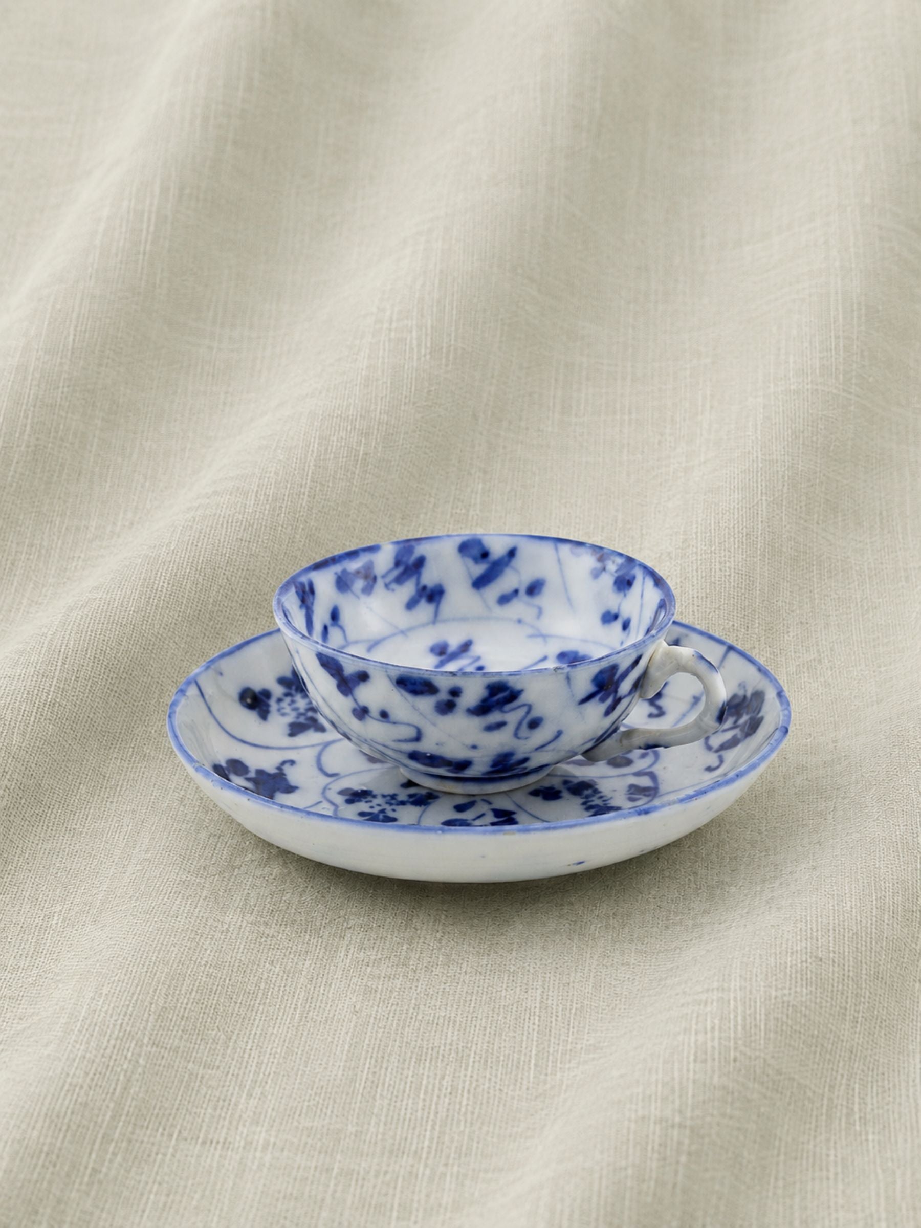 A small white ceramic teacup and saucer set decorated with a blue floral pattern, placed on a textured beige fabric background.