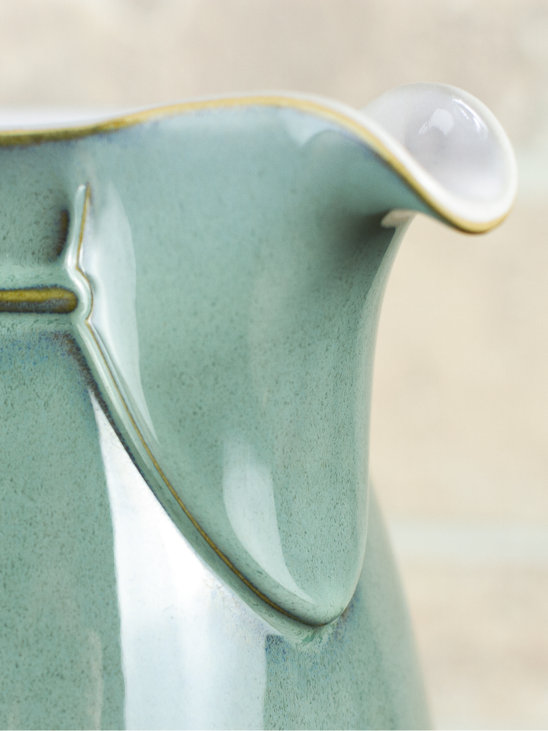 Close-up of a glossy ceramic pitcher with a pale sage green glaze and thin gold trim, showing the spout and handle against a softly blurred neutral background.