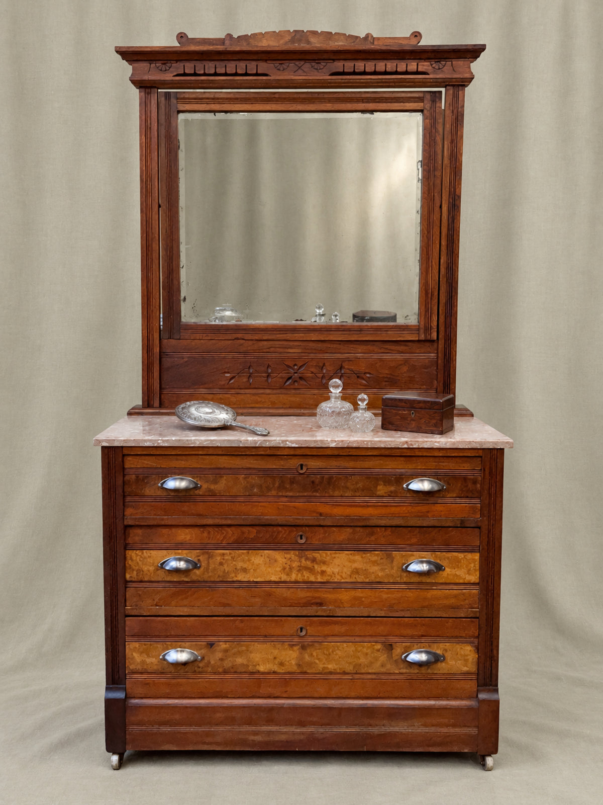 An antique Eastlake wooden dresser with a large mirror, featuring multiple drawers with silver handles and a light-colored marble top, set against a neutral draped backdrop.