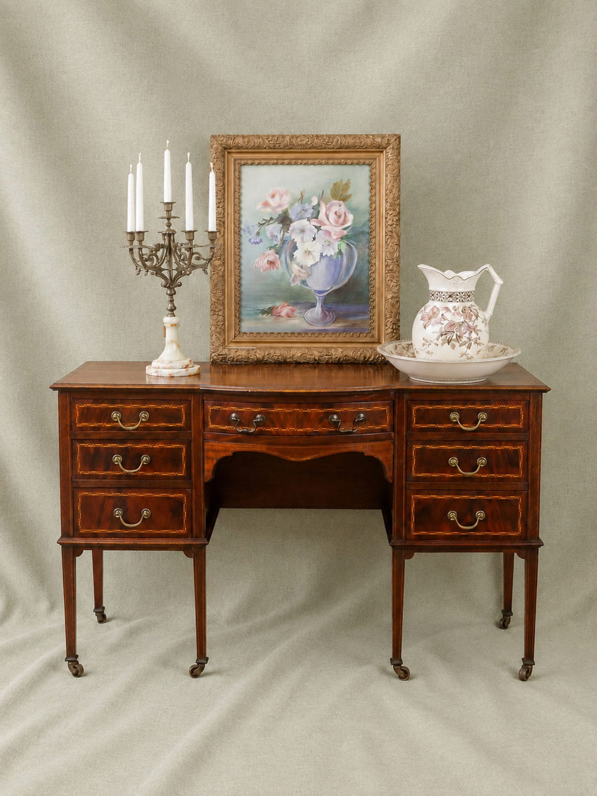 A vintage mahogany writing desk with ornate drawer pulls, styled with a framed floral painting, a white candelabra, and a white floral pitcher on a draped beige fabric background.