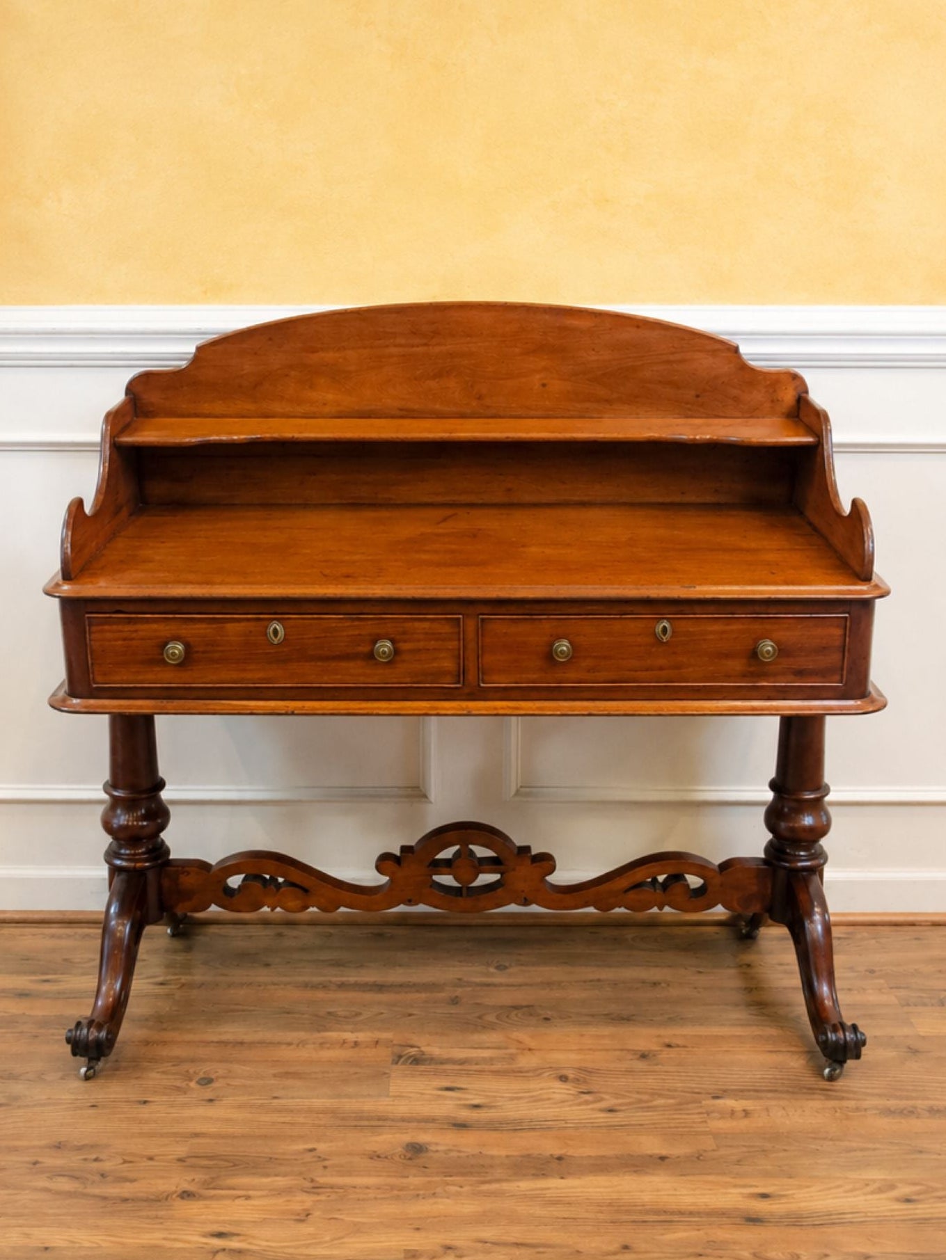 An antique wooden writing desk with a curved backrest, two drawers, and ornate turned legs, set against a yellow and white paneled wall on a wooden floor.