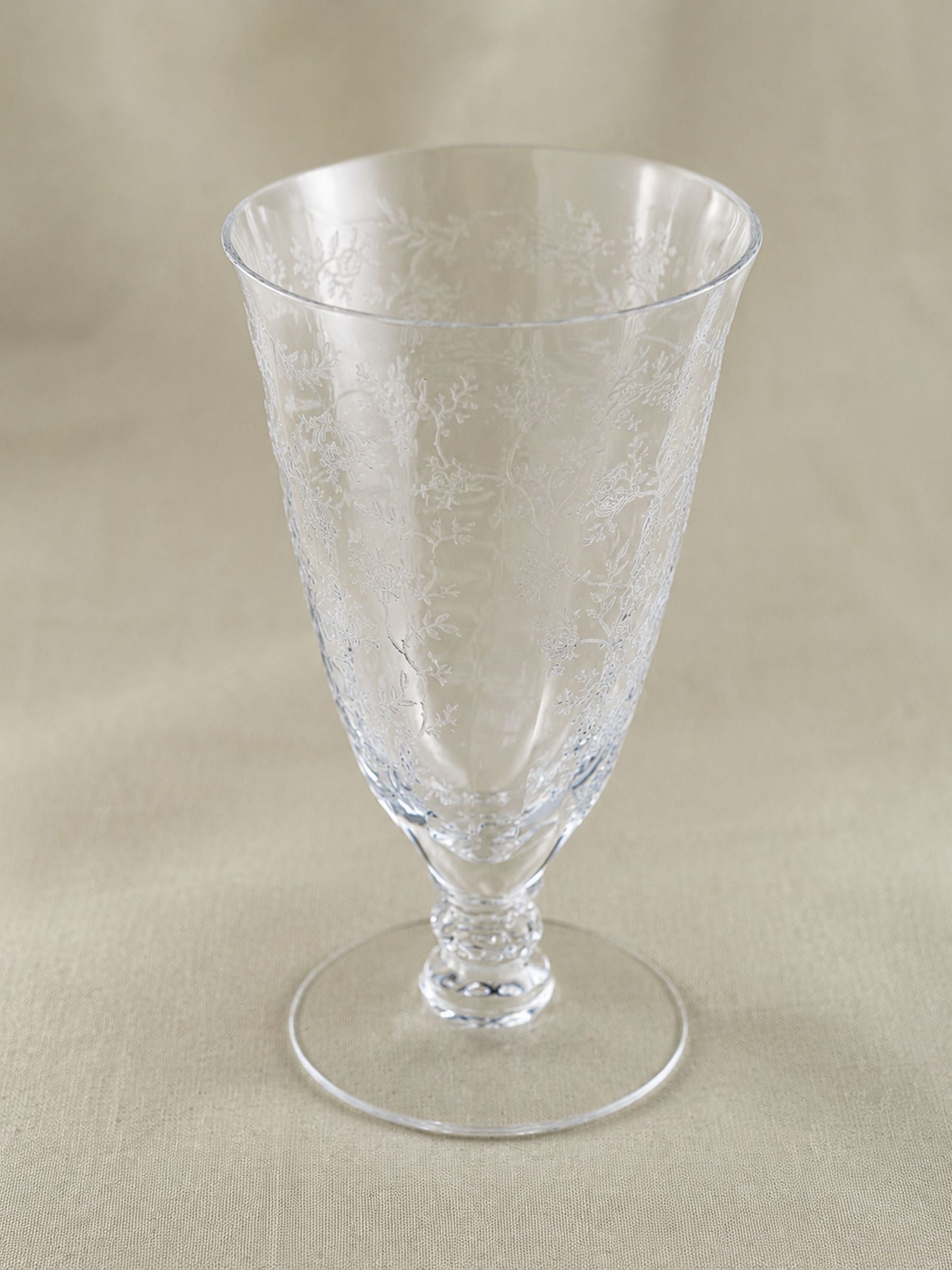 A clear glass goblet with delicate etched floral patterns, set against a neutral beige fabric background.