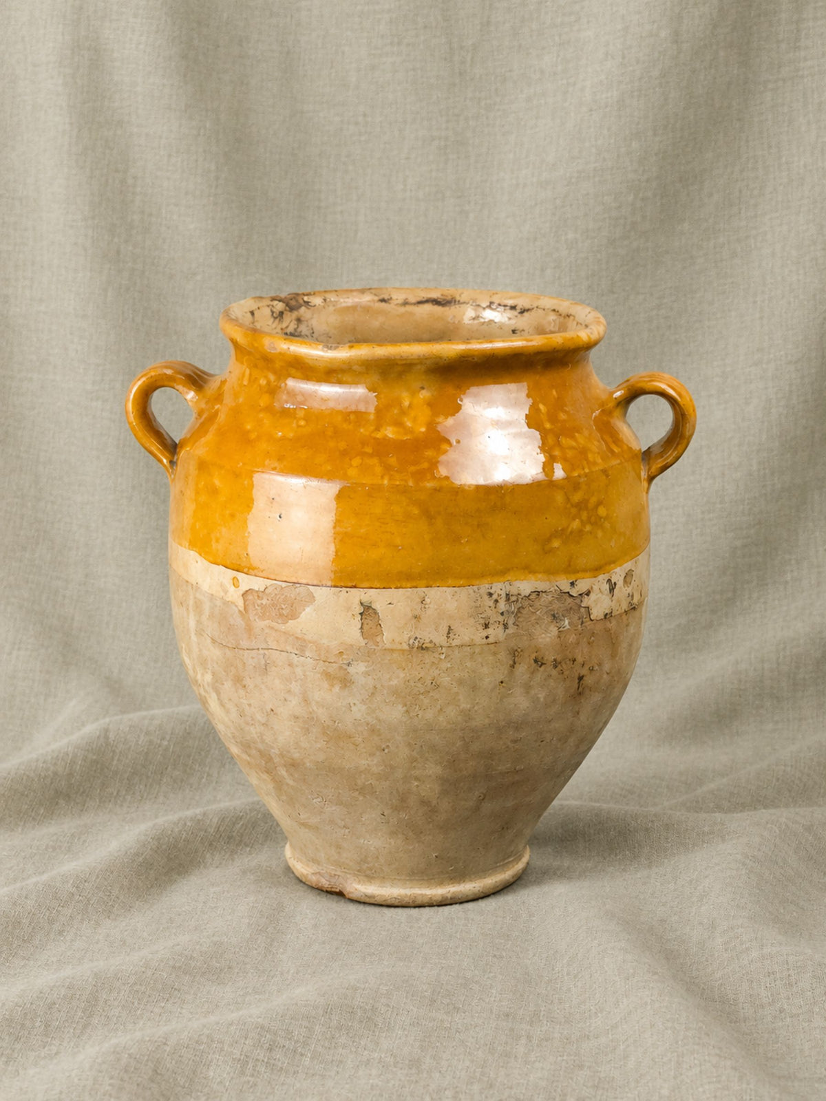 A French antique ceramic vase with a two-tone glaze, featuring a glossy ochre yellow upper section and a textured beige lower section, set against a draped linen backdrop.