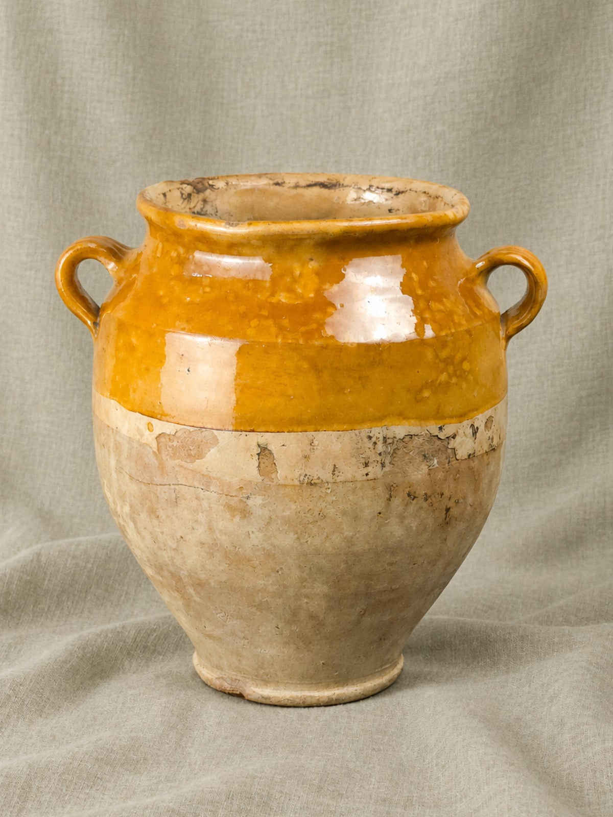 A French antique ceramic confit pot with a two-tone glaze, featuring a glossy ochre yellow upper section and a textured beige lower section, set against a draped linen backdrop.