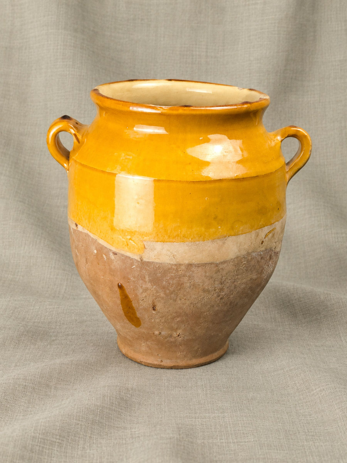 A handmade ceramic confit pot with a two-tone glaze, featuring a bright yellow upper section and a natural brown lower section, set against a neutral gray fabric backdrop.