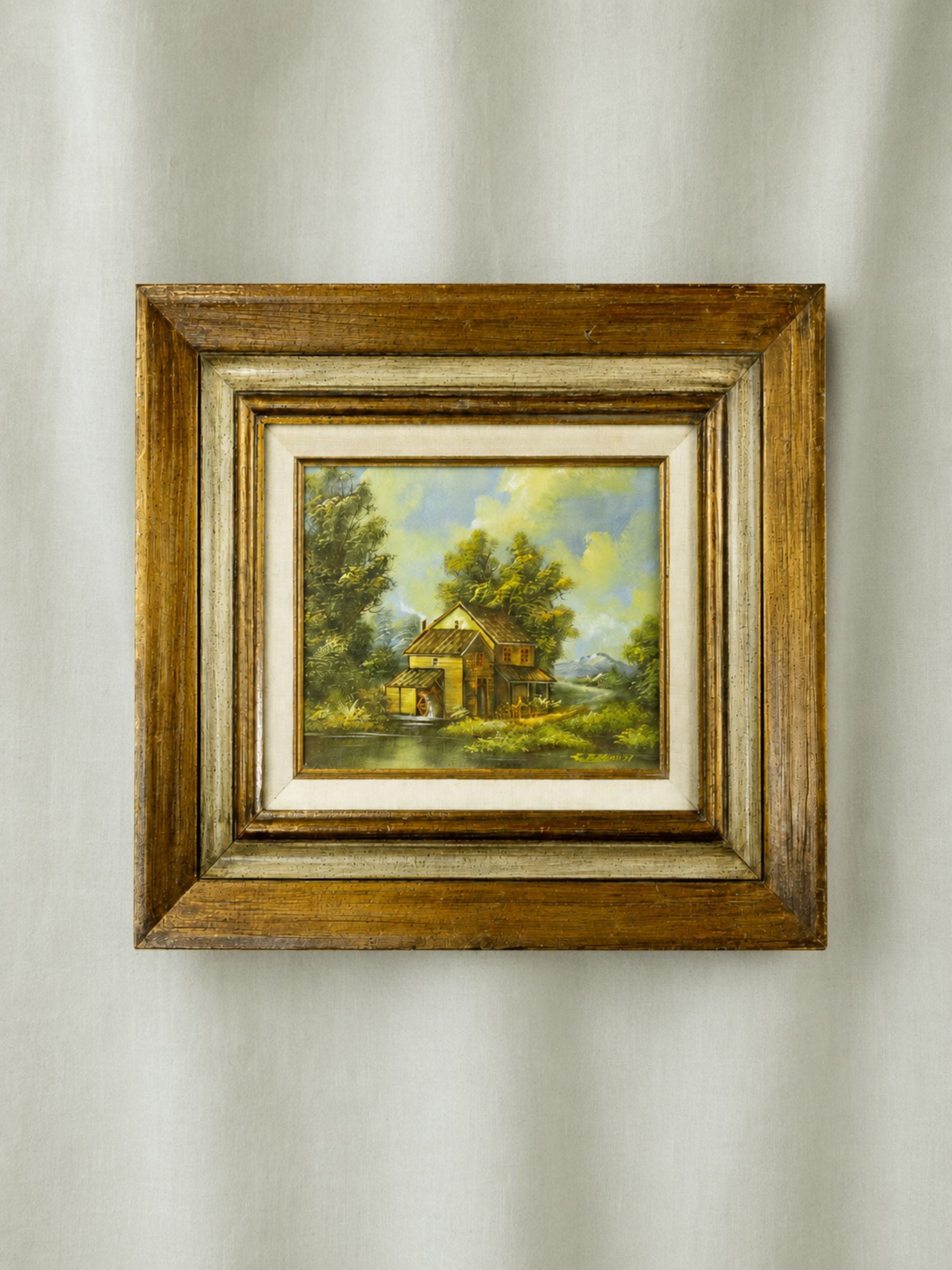 A small framed landscape painting of a rustic yellow house surrounded by green trees under a blue sky, set in a distressed brown wooden frame with a cream mat, hanging on a light gray fabric wall.