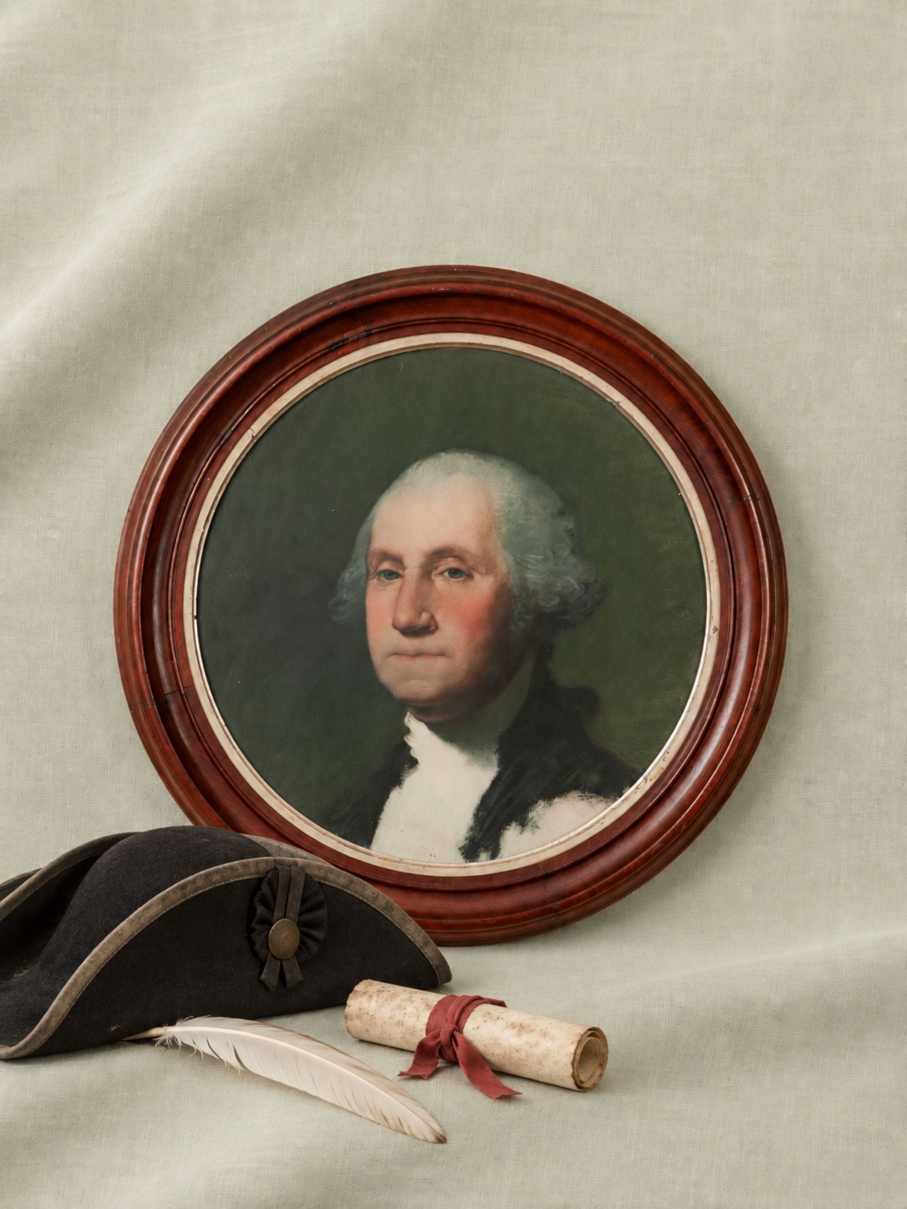 A framed portrait of George Washington on a draped fabric background, styled with a black tricorn hat, a white feather quill, and a rolled paper document tied with a red ribbon.