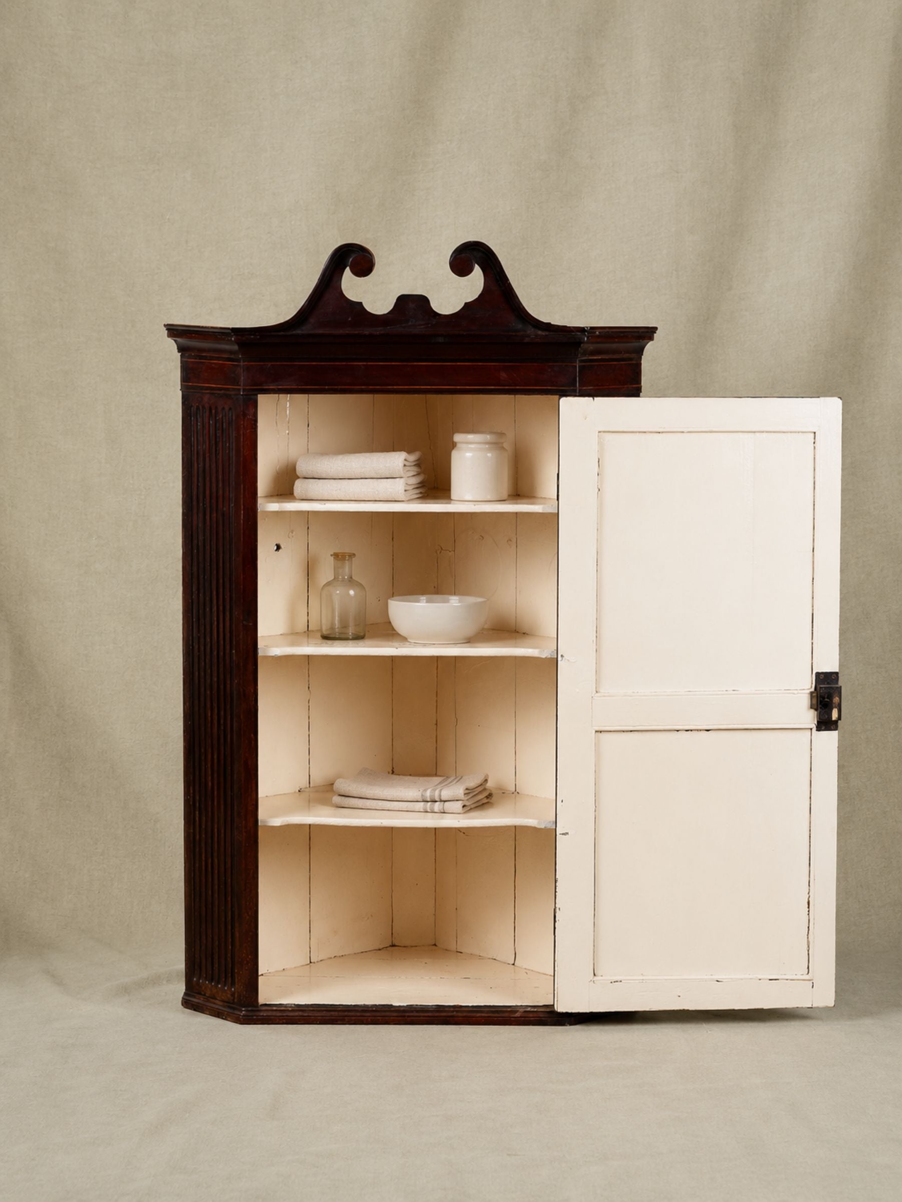 A antique corner cabinet with a dark brown wood frame and a distressed off-white door, shown open with shelves holding white dishes against a neutral beige backdrop.