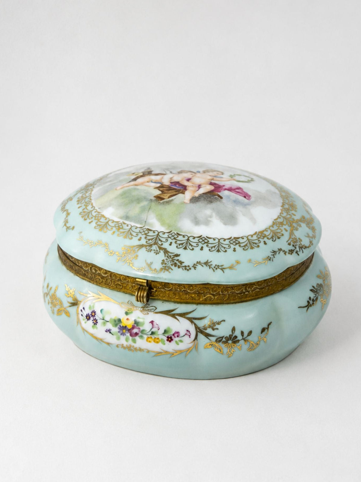 A round, light aqua porcelain trinket box with a lid, decorated with gold floral patterns, a gold-tone clasp, and hand-painted floral details on the lid and front.