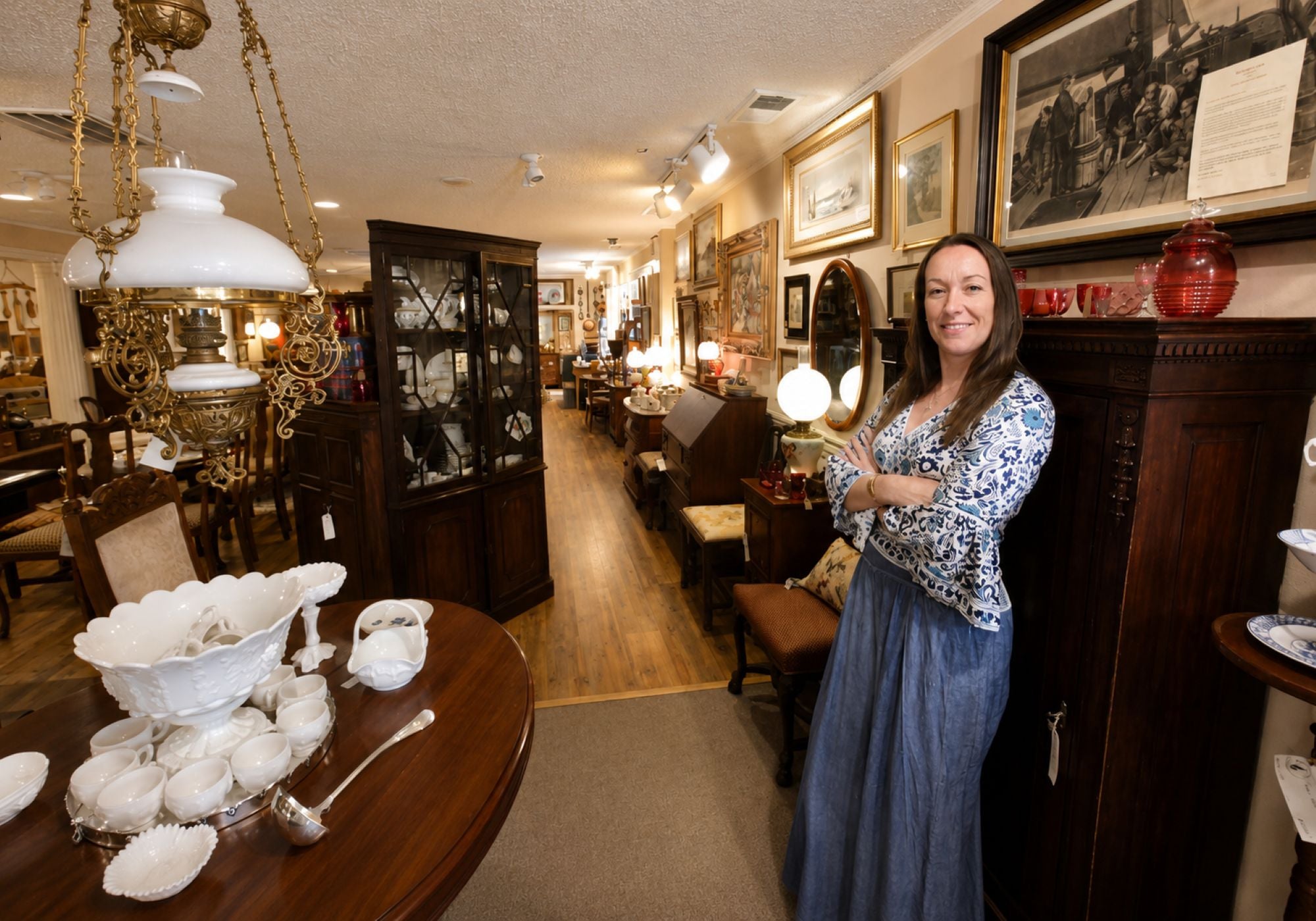 A woman stands in a vintage furniture and home decor showroom, smiling next to a dark wood cabinet. The room is filled with antique lamps, framed pictures, and other furniture pieces, with a round wooden table in the foreground holding a white ceramic bowl and silverware.