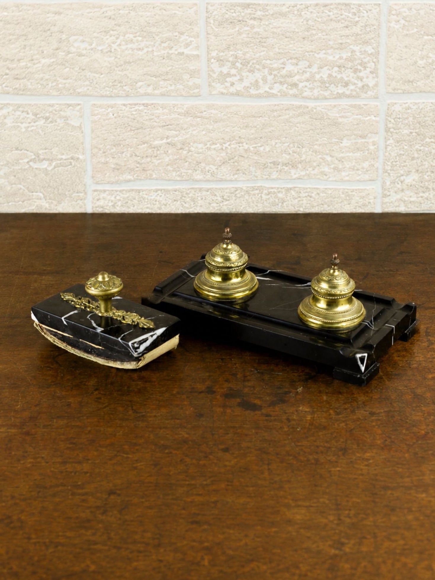 A vintage black marble desk set with gold brass inkwells and a matching gold handled ink blotter, displayed on a dark wooden table against a light brick wall.