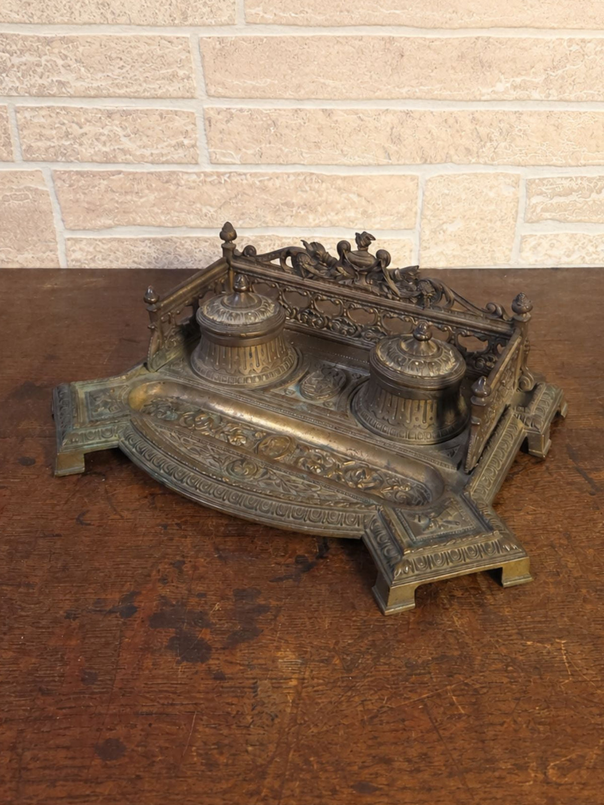 An ornate antique bronze desk inkwell, with 2 lidded wells and decorative scrollwork, displayed on a dark wooden table against a light brick wall.