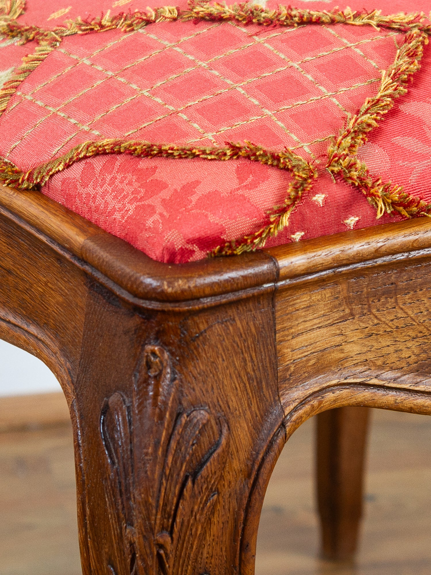 Close-up of a carved wooden chair leg and seat upholstered in red fabric with a gold diamond lattice pattern and gold fringe trim, set against a warm wood floor background.