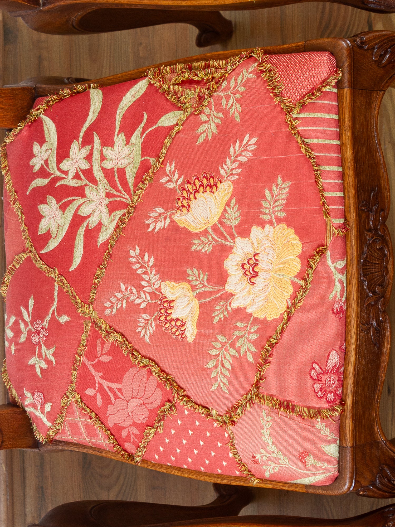 A close-up of a decorative upholstered chair seat with a patchwork design in shades of coral pink, red, and peach, featuring cream floral motifs and gold fringe trim, set against a dark carved wooden frame.