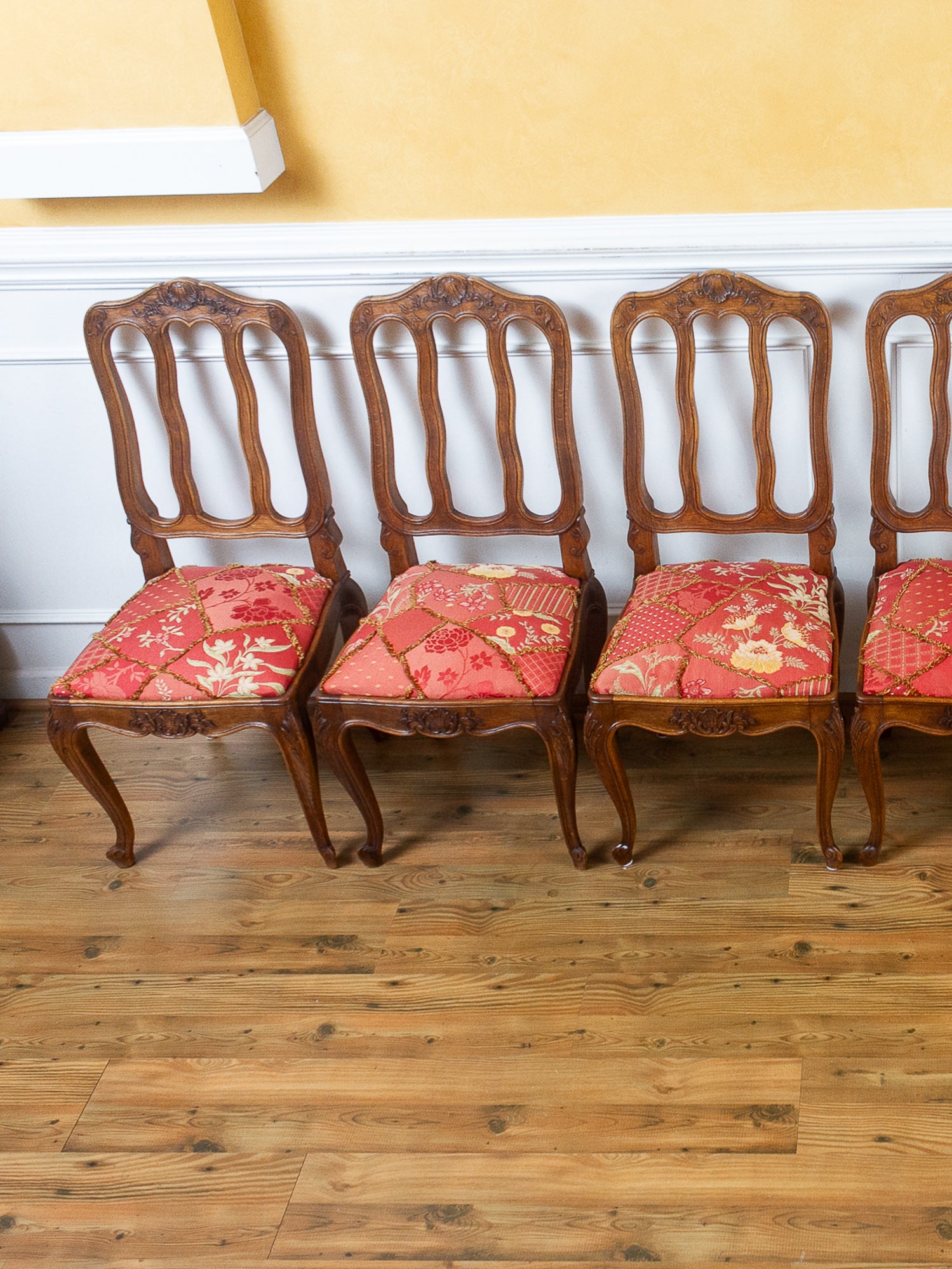 Four antique wooden dining chairs with carved backs and red floral upholstered seats, arranged on a hardwood floor against a yellow wall.