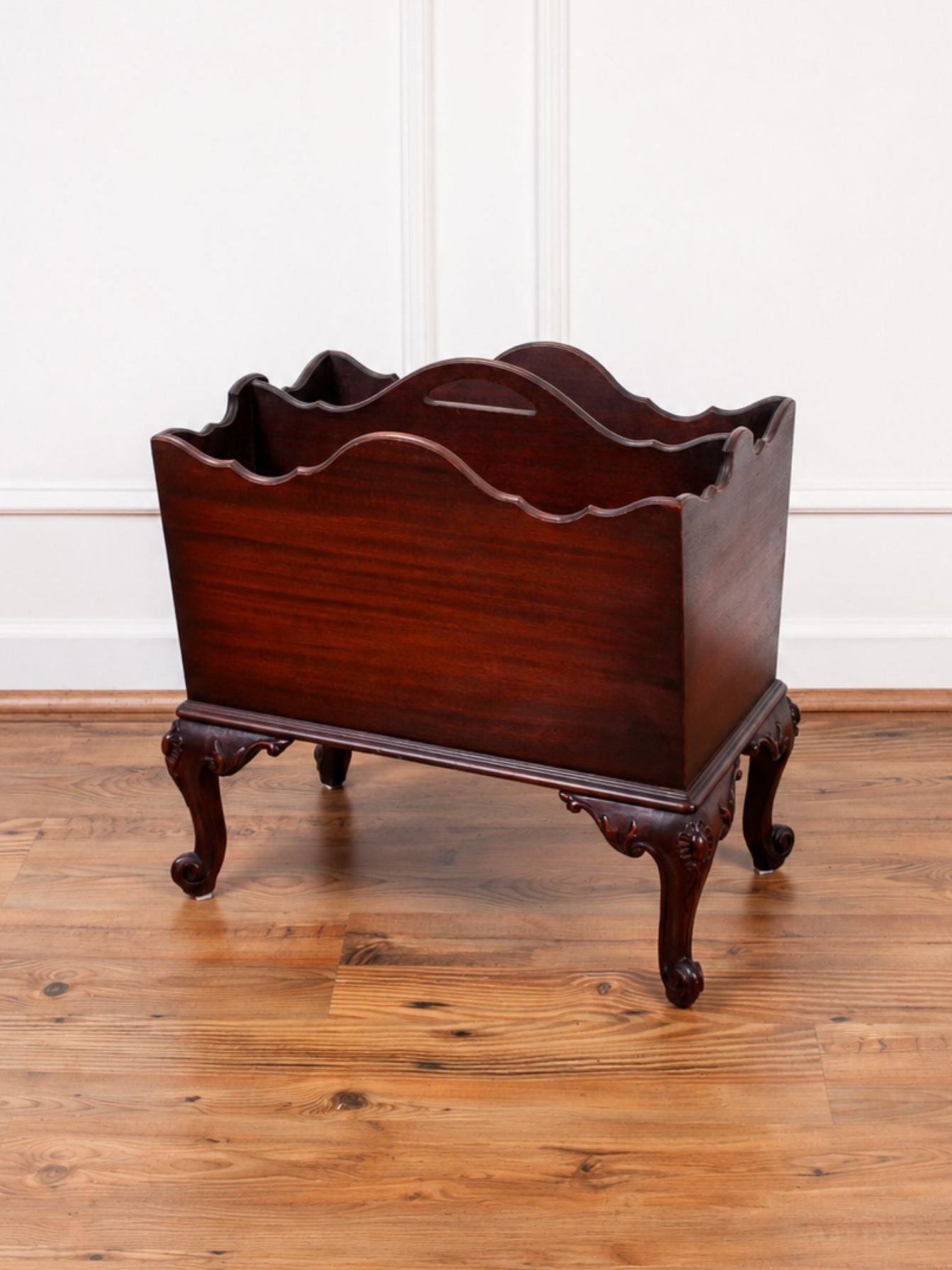 A dark reddish-brown mahogany wooden magazine rack with carved legs, shown on a polished wood floor against a white paneled wall.