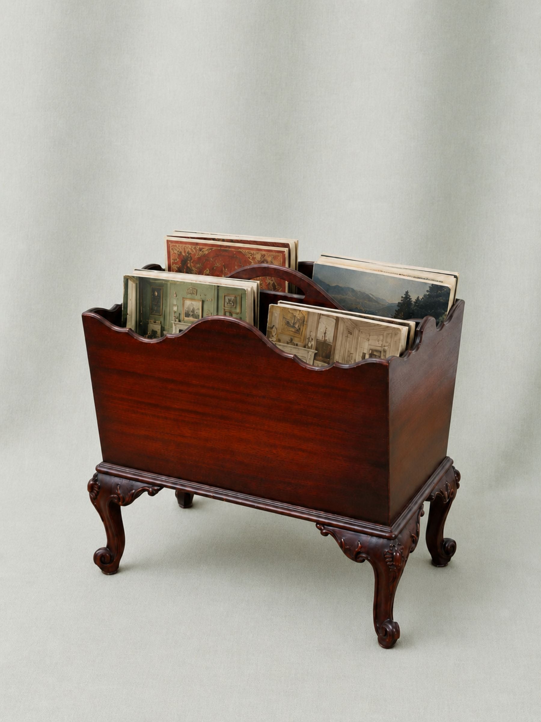 A vintage-style wooden magazine rack with carved cabriole legs and a scalloped front edge, holding several printed magazines or booklets, photographed against a neutral draped backdrop.
