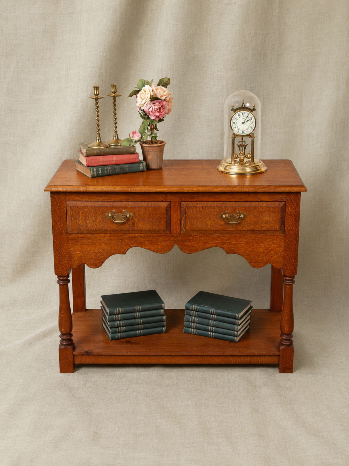 A antique oak wooden console table with two drawers and a lower shelf, styled with books, a vase of flowers, candlesticks, and a glass-domed clock against a beige fabric backdrop.