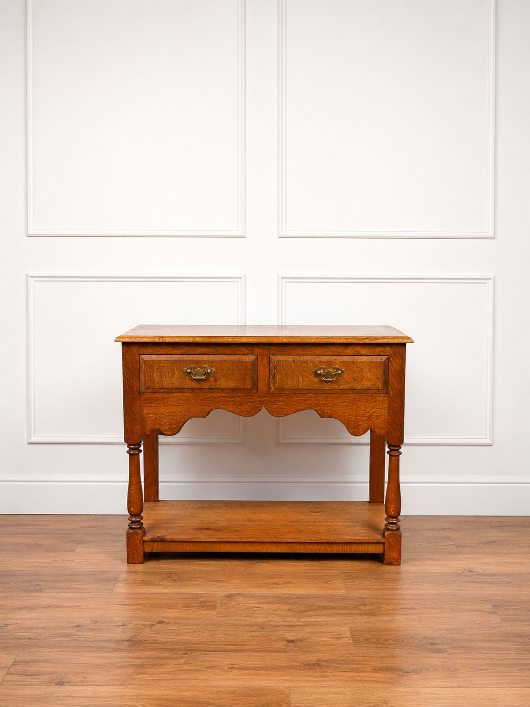 An antique oak wooden console table with two drawers, turned legs, and a lower shelf, set against a white paneled wall and hardwood floor.