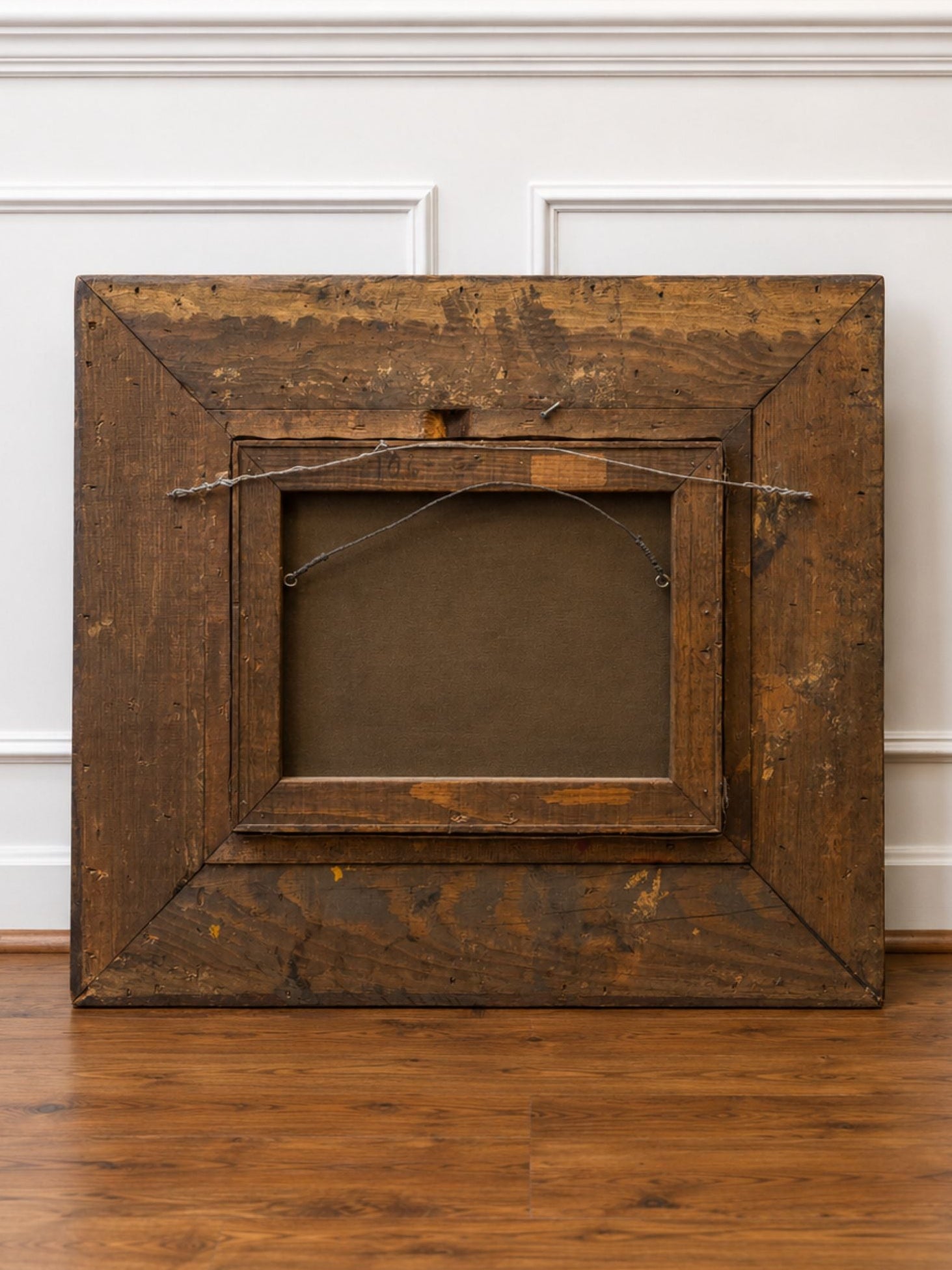 A large, rustic antique wooden picture frame with a dark brown finish, shown from the back with its hanging wire, leaning against a white paneled wall on a hardwood floor.