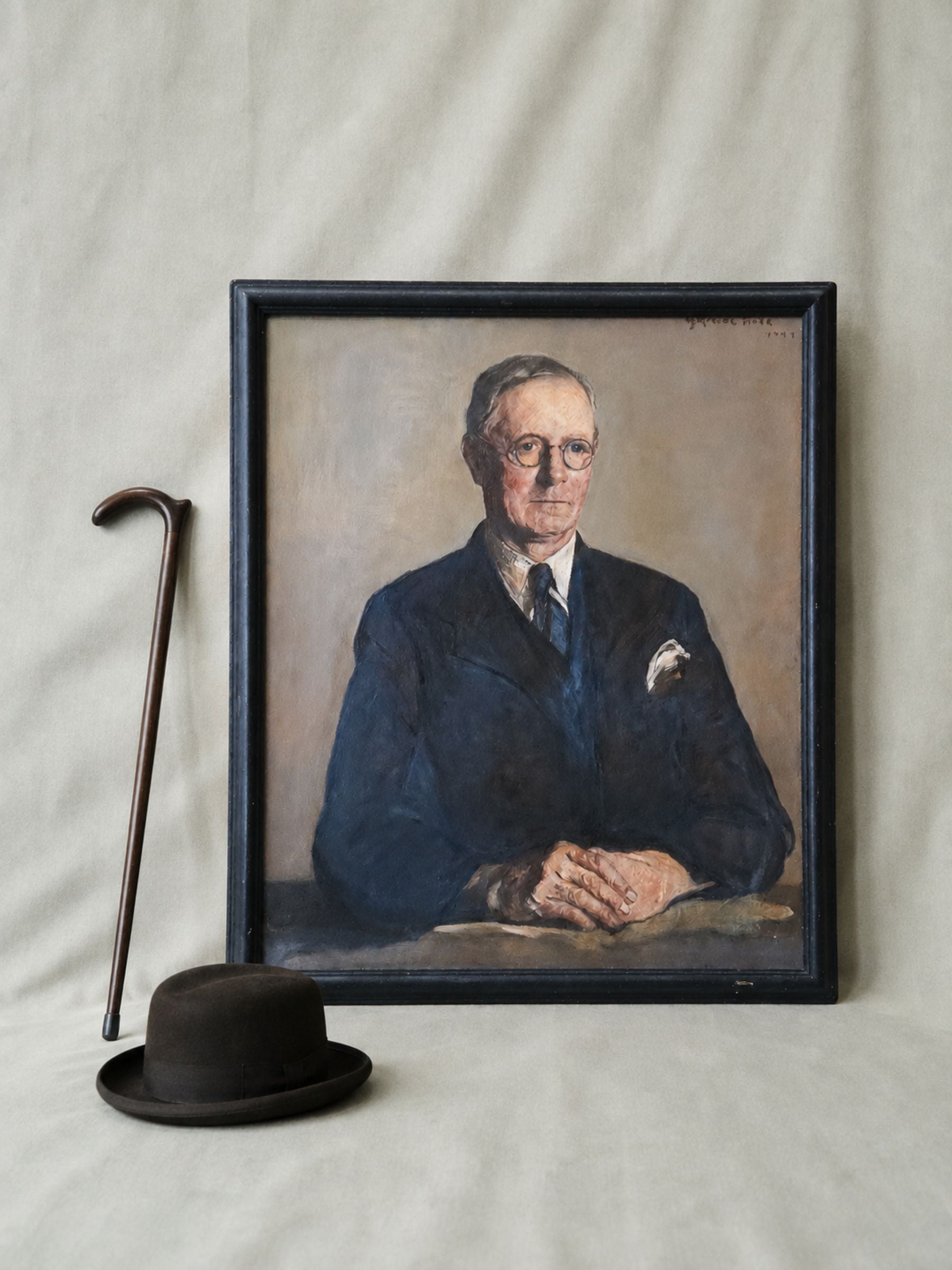 A framed portrait painting by Gertrude Horsford Fiske of a man in a dark suit, displayed against a draped fabric backdrop, with a cane and a black bowler hat placed beside it.