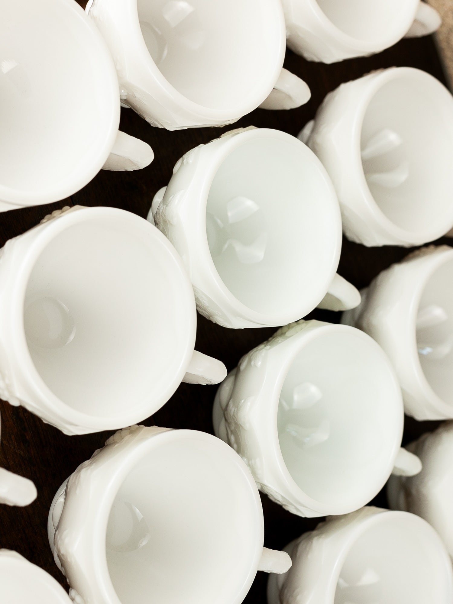 A close-up, top-down view of multiple white milk glass teacups arranged closely together on a dark surface.