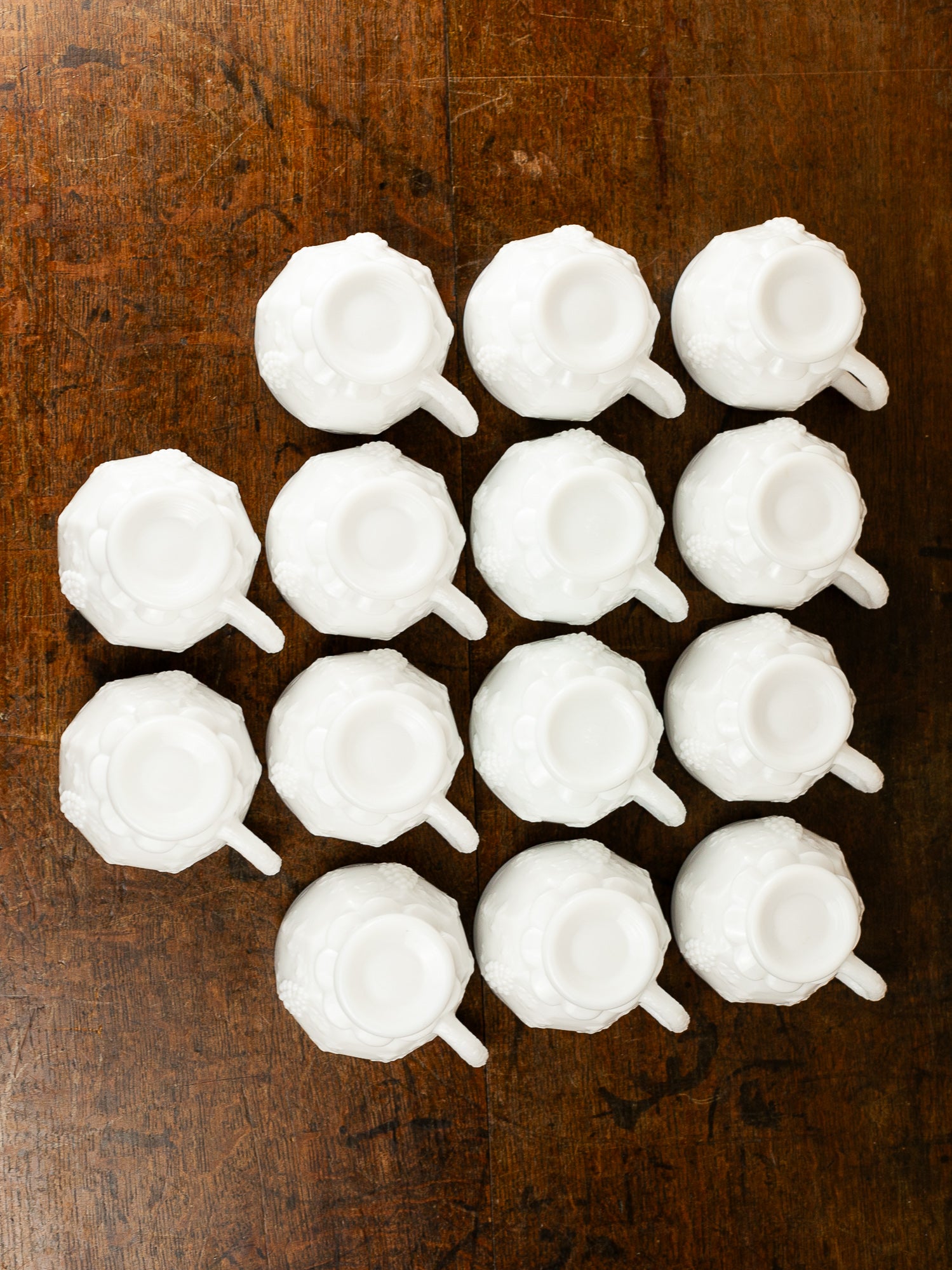 Fourteen small milk glass teacups arranged in a grid on a dark brown wooden tabletop, photographed from above showing the under sides.