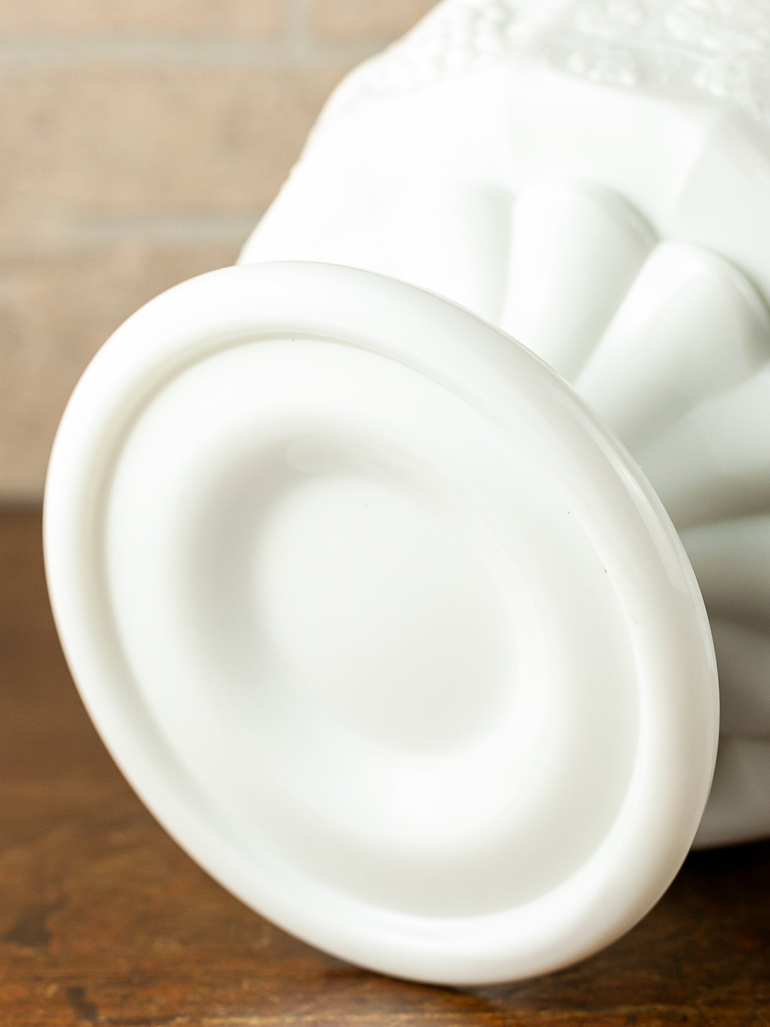 Close-up of a white milk glass pedestal base showing its flared circular base, resting on a dark wooden surface with a blurred beige brick wall in the background.