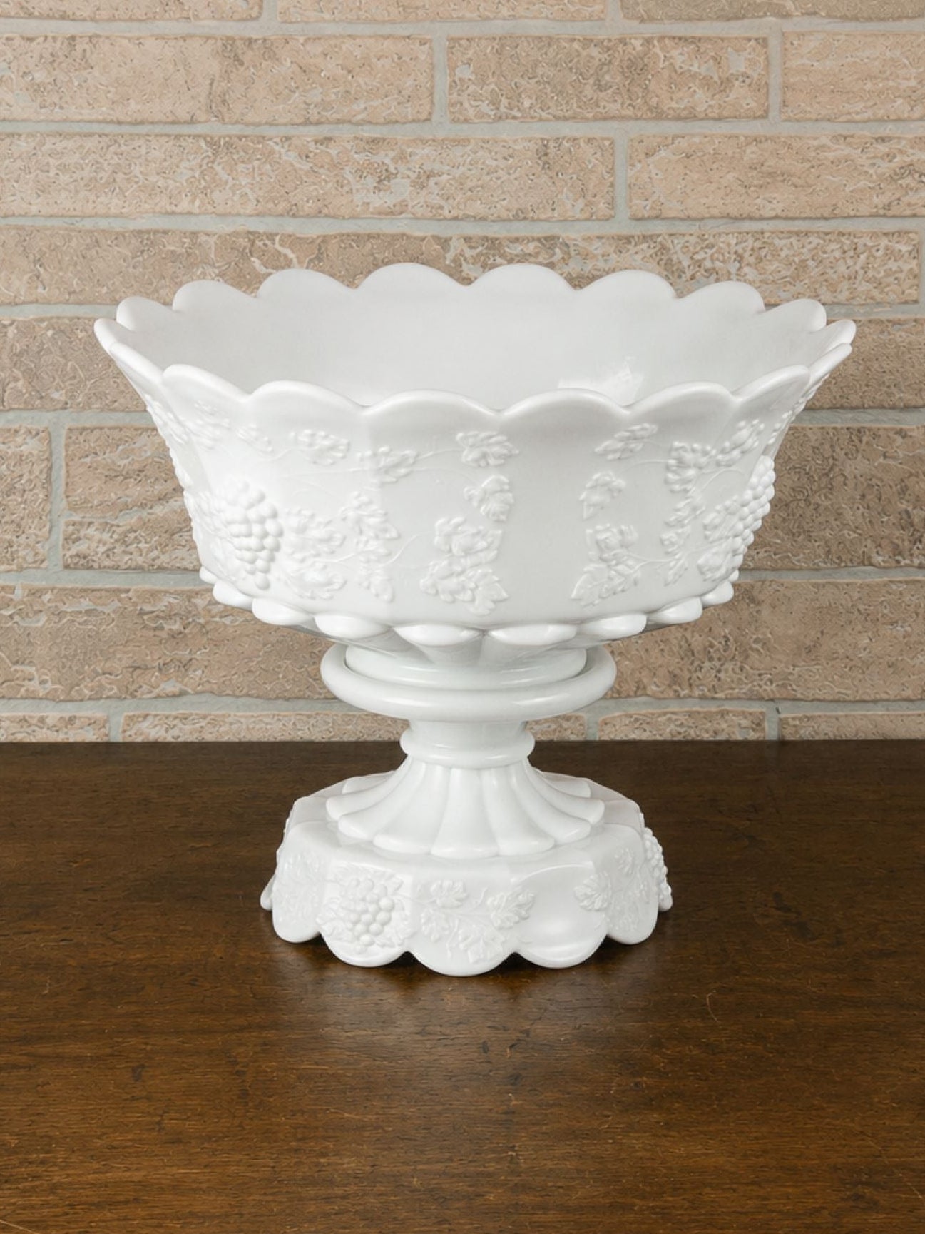 A white glass pedestal bowl with a scalloped rim and embossed grapevine pattern, set on a dark wooden table against a light brick wall.