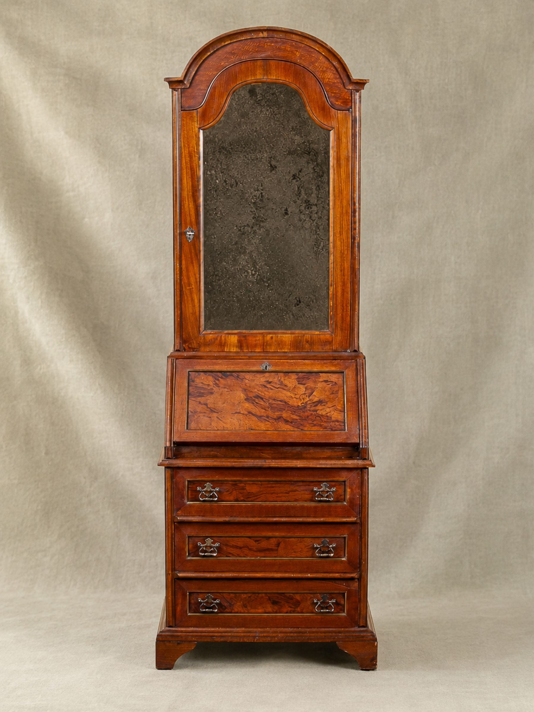 An antique mahogany dresser/writing desk with an attached mirror and three drawers, set against a neutral beige fabric backdrop.