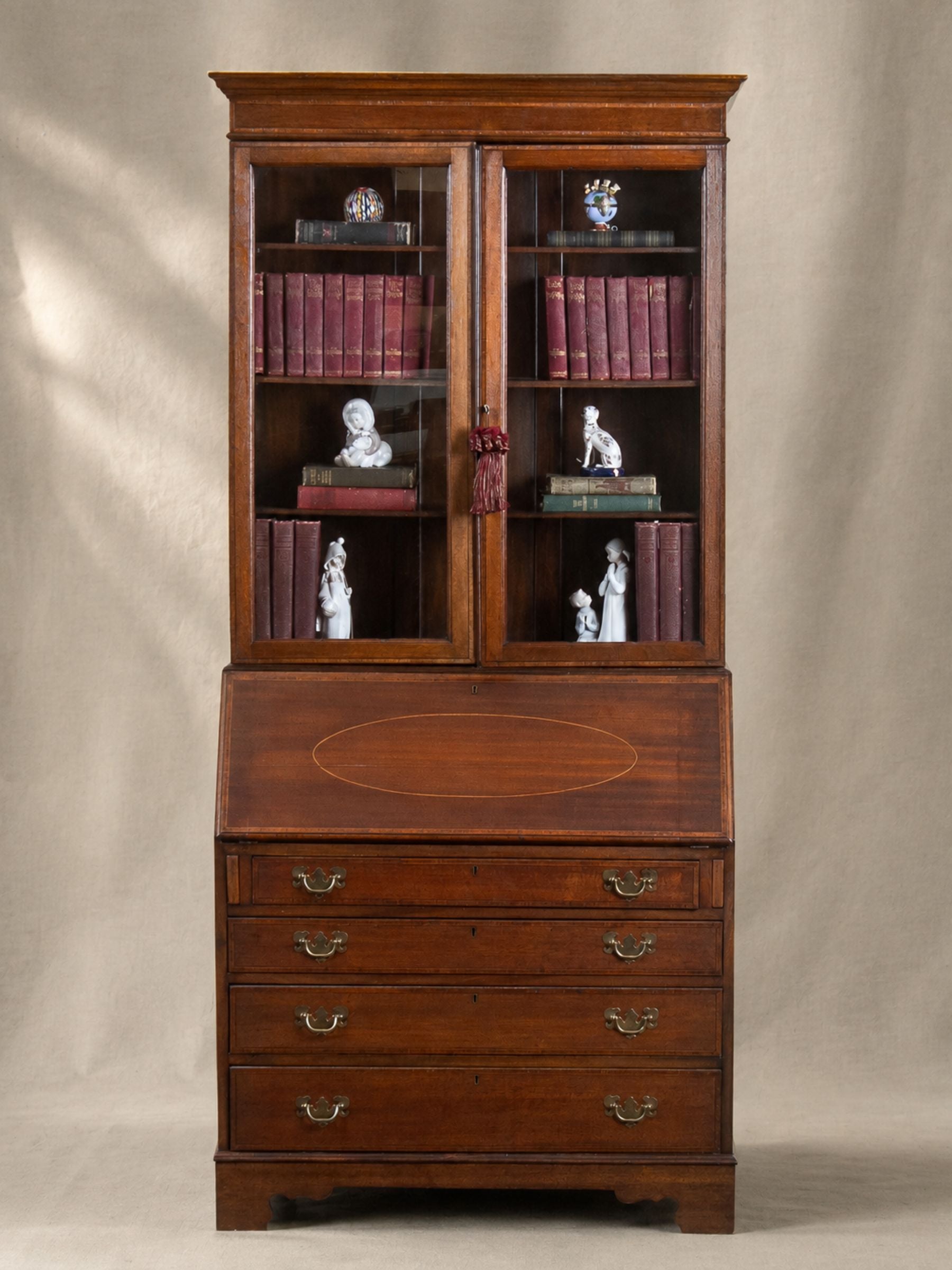 A tall antique mahogany writing desk with a hutch, featuring glass-paneled upper doors, decorative bookshelves with books and figurines, a writing surface with an oval inlay, and five drawers below with brass handles, set against a neutral beige backdrop.
