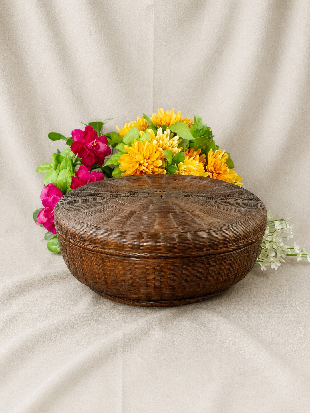 A round, dark brown woven wicker basket with a fitted lid, styled with artificial yellow and pink flowers on a draped beige fabric background.